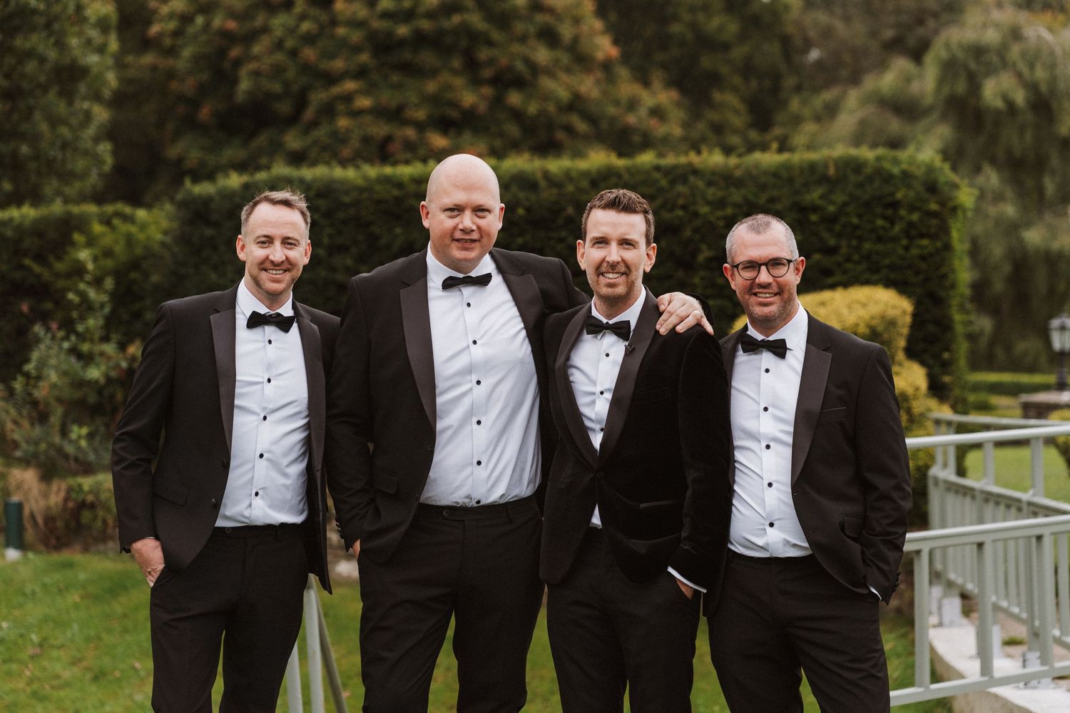 Groomsmen in black tuxedos and bow ties pose together outdoors at an elegant wedding celebration.