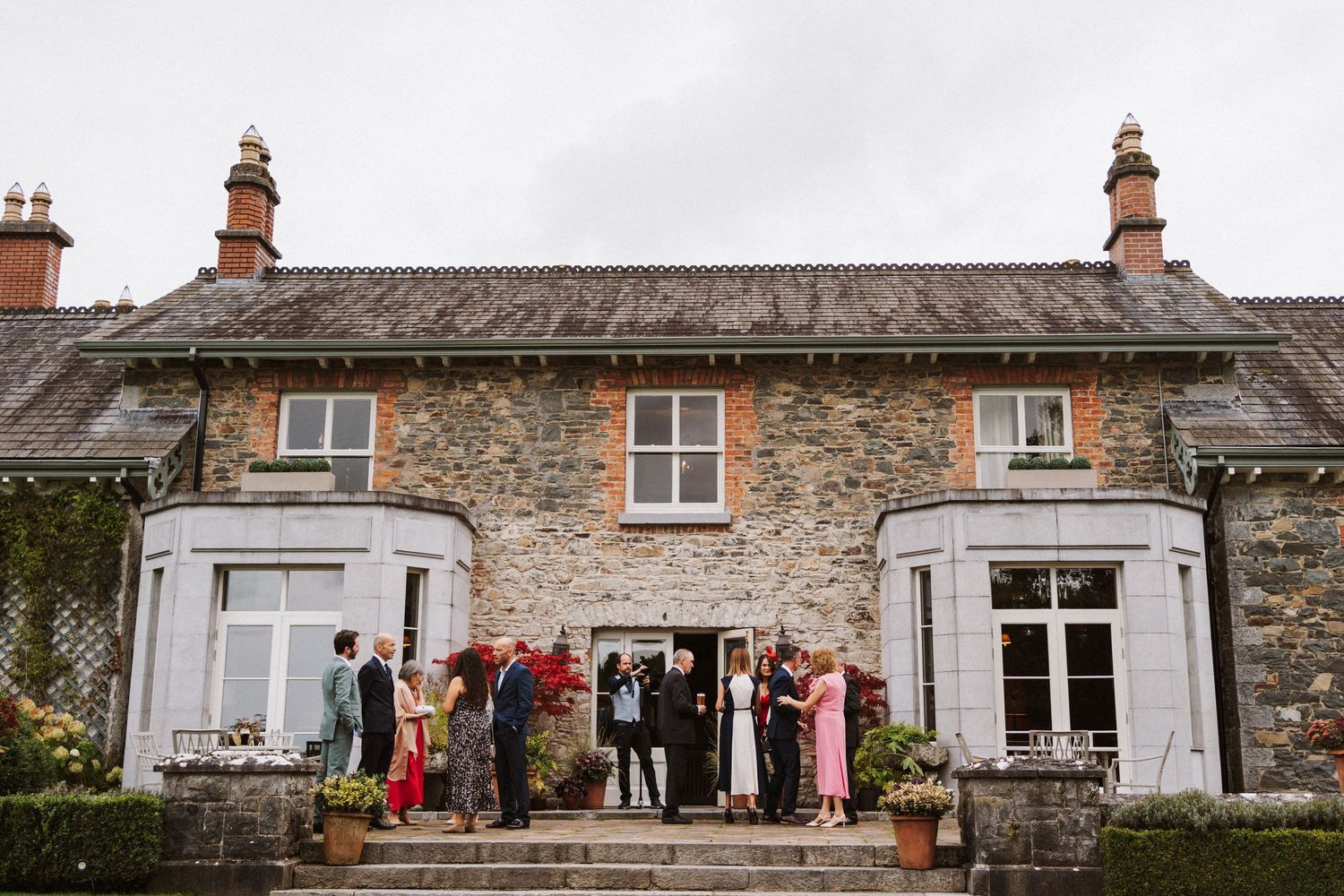 Wedding guests stand outside a rustic stone cottage with white bay windows and red accents during a celebration.