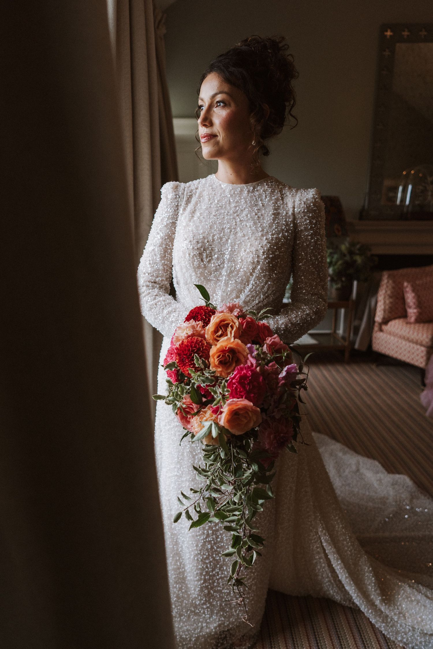Bride in lace dress holding vibrant coral and burgundy floral bouquet near window light.