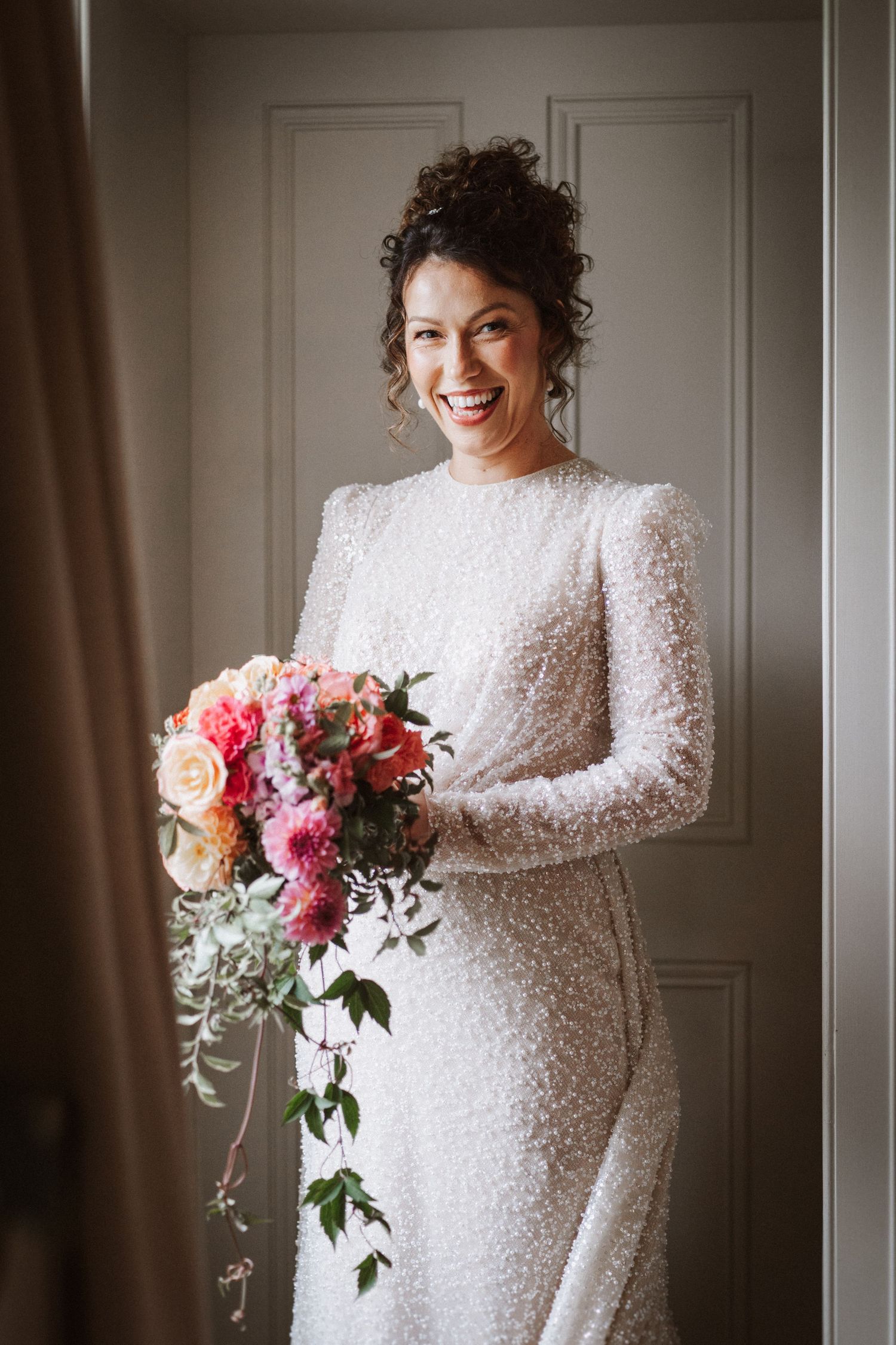Elegant bridal portrait featuring a long-sleeved beaded white gown and vibrant pink and peach floral bouquet.