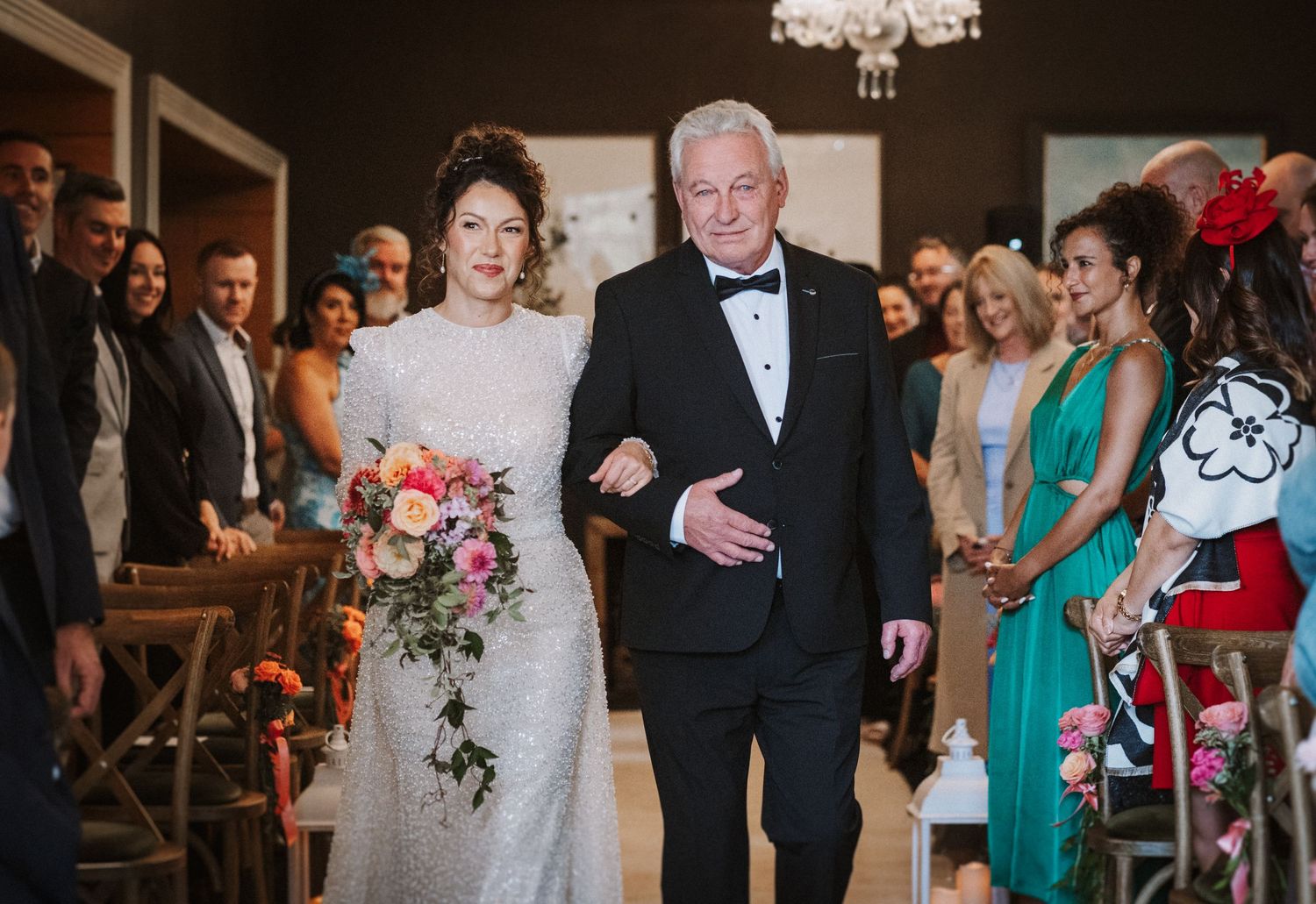 Wedding guests in formal attire gather at an elegant reception venue with a crystal chandelier overhead.