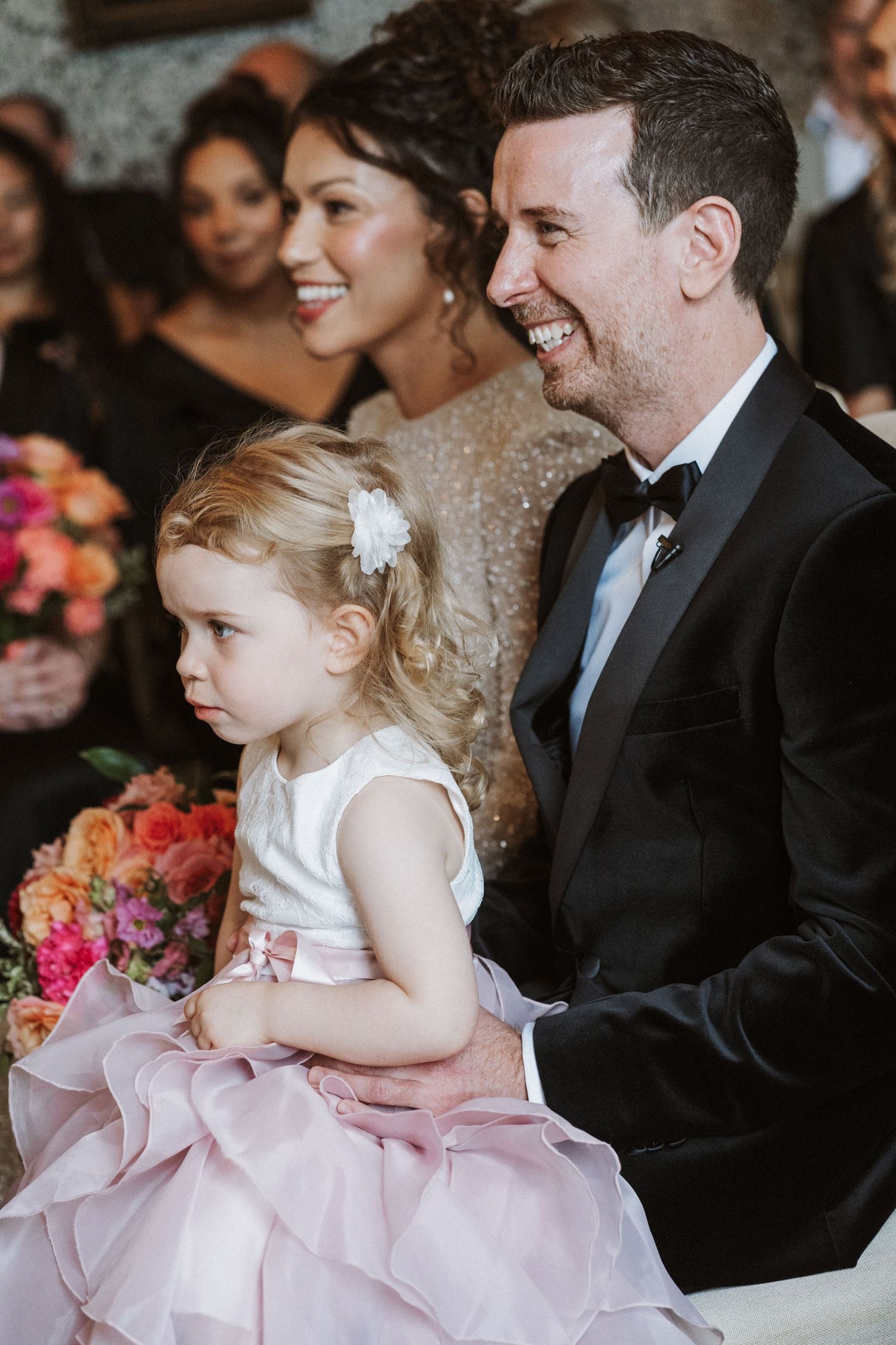 A young flower girl in a pink dress sits among formally dressed guests at a wedding ceremony.