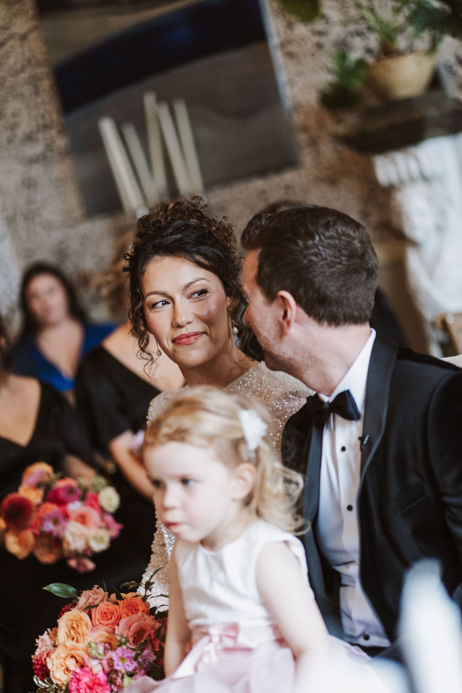 Wedding ceremony attendees gather in an elegant room with ornate wallpaper and colorful floral arrangements.