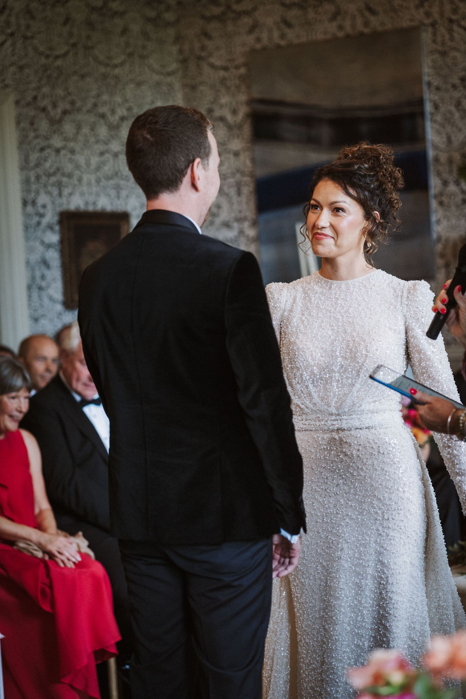Wedding ceremony in progress at an elegant venue with damask wallpaper and formal attire.
