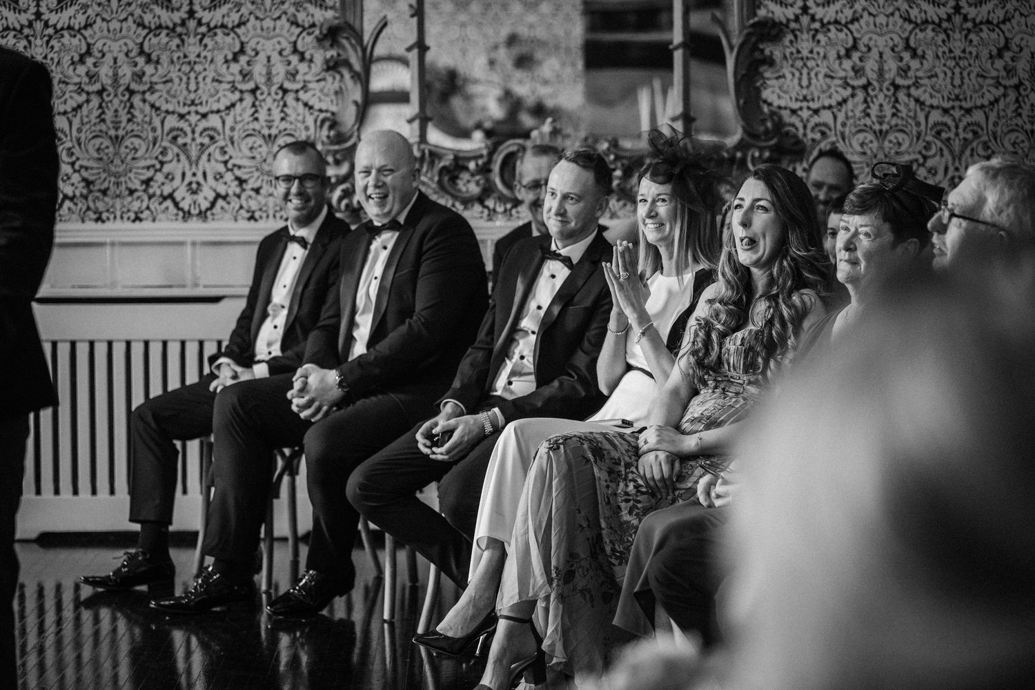Wedding guests seated in formal attire against an elegant wallpapered backdrop.