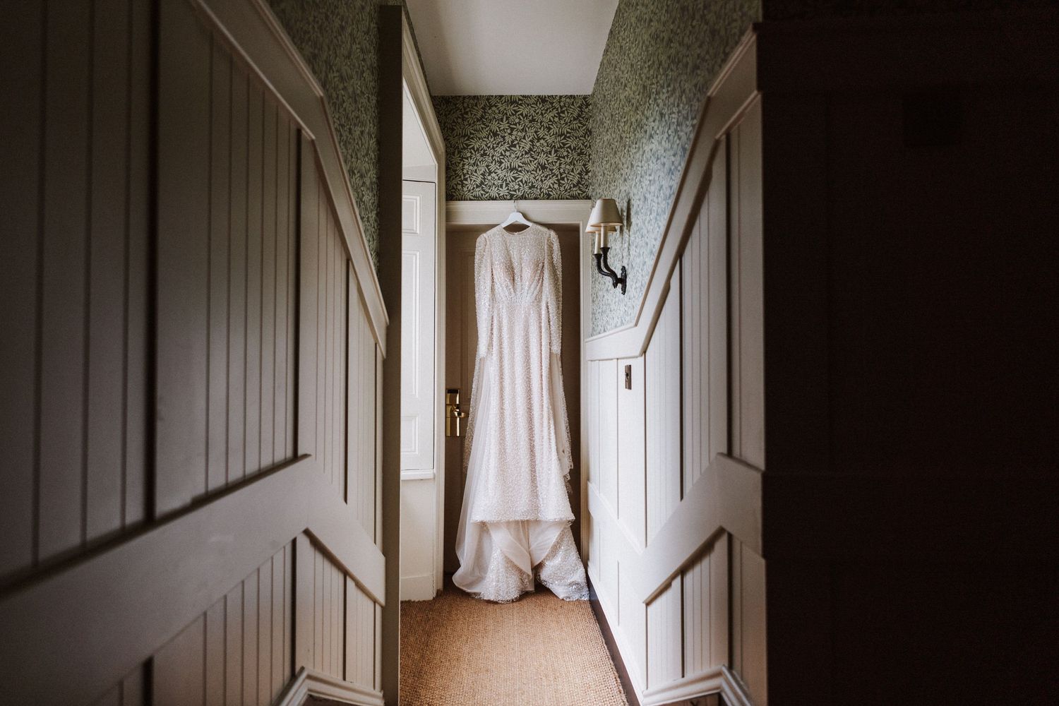 White wedding dress hanging in a dark wooden doorway creating a dramatic silhouette against natural light.
