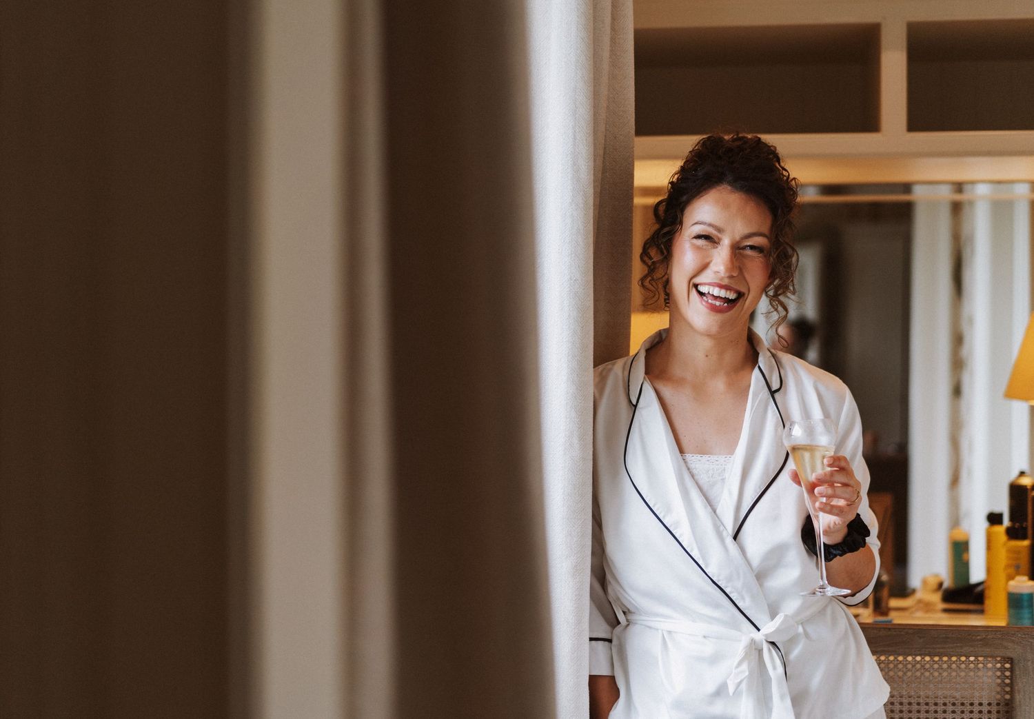 A person in a white robe stands in a sunlit room smiling and laughing while holding a wine glass.