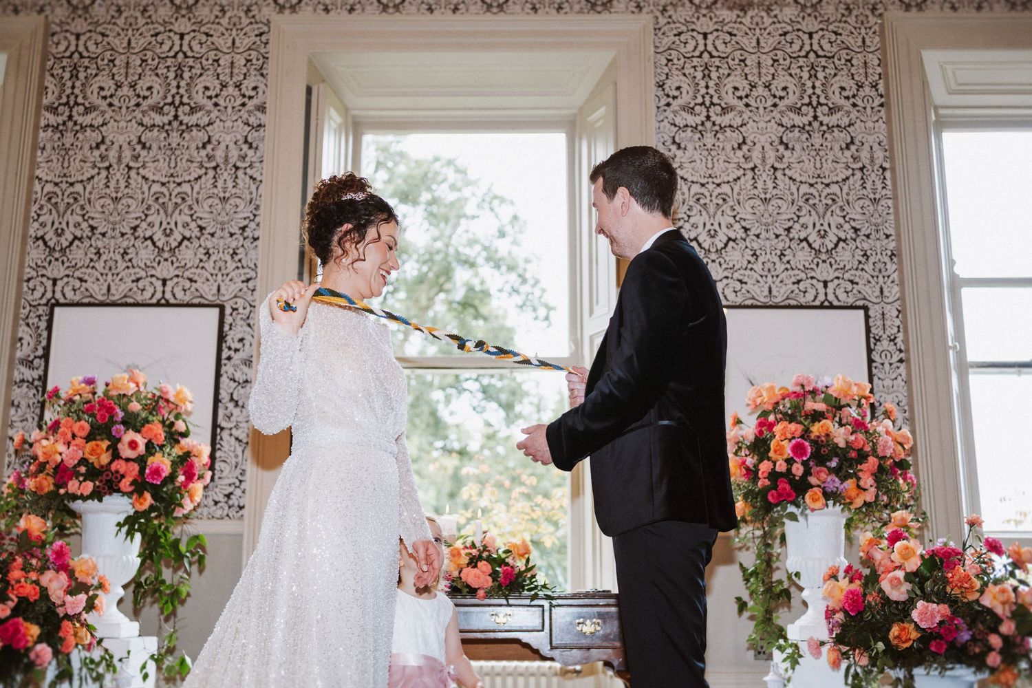 Wedding ceremony in an elegant room with floral wallpaper and bright flower arrangements.
