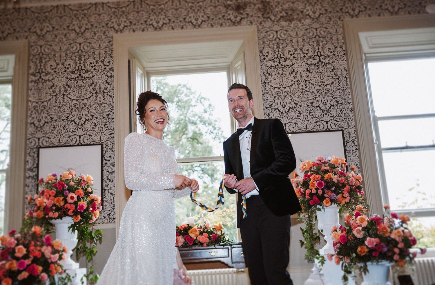 Wedding ceremony in a room with ornate wallpaper and bright floral arrangements by windows.