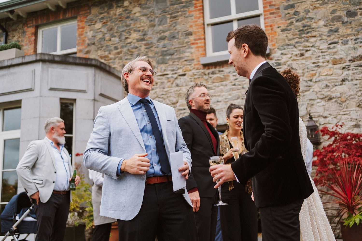 A group of wedding guests share joyful laughter outside a rustic stone building during a celebration.