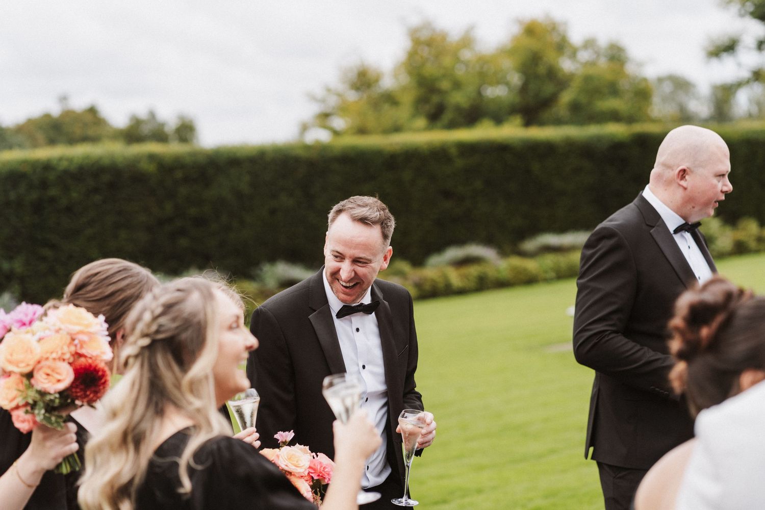 Wedding guests gather outdoors on a lush green lawn during a sunny celebration at an elegant venue.