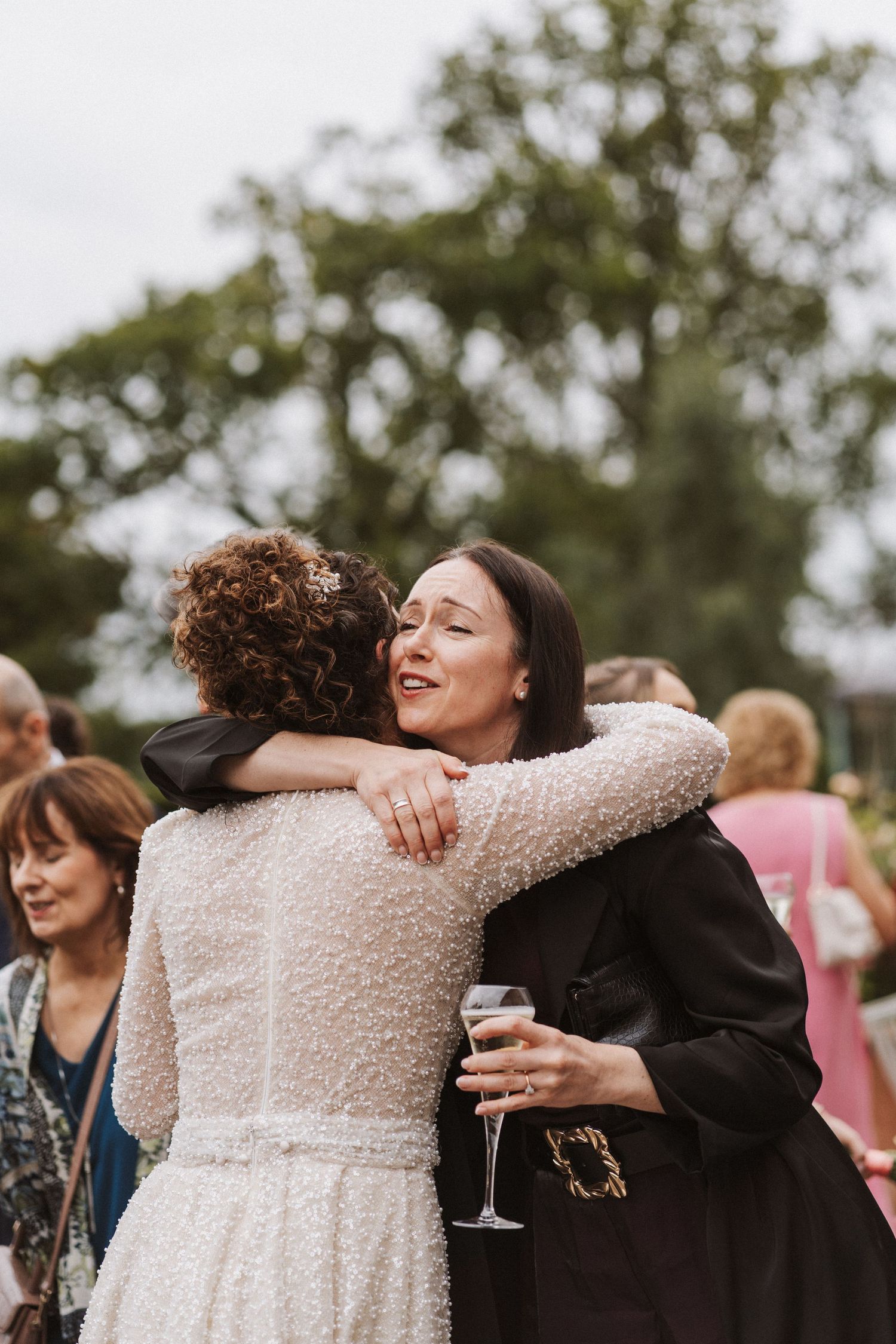 Two wedding guests share a warm embrace at an outdoor celebration while one holds a glass of champagne.
