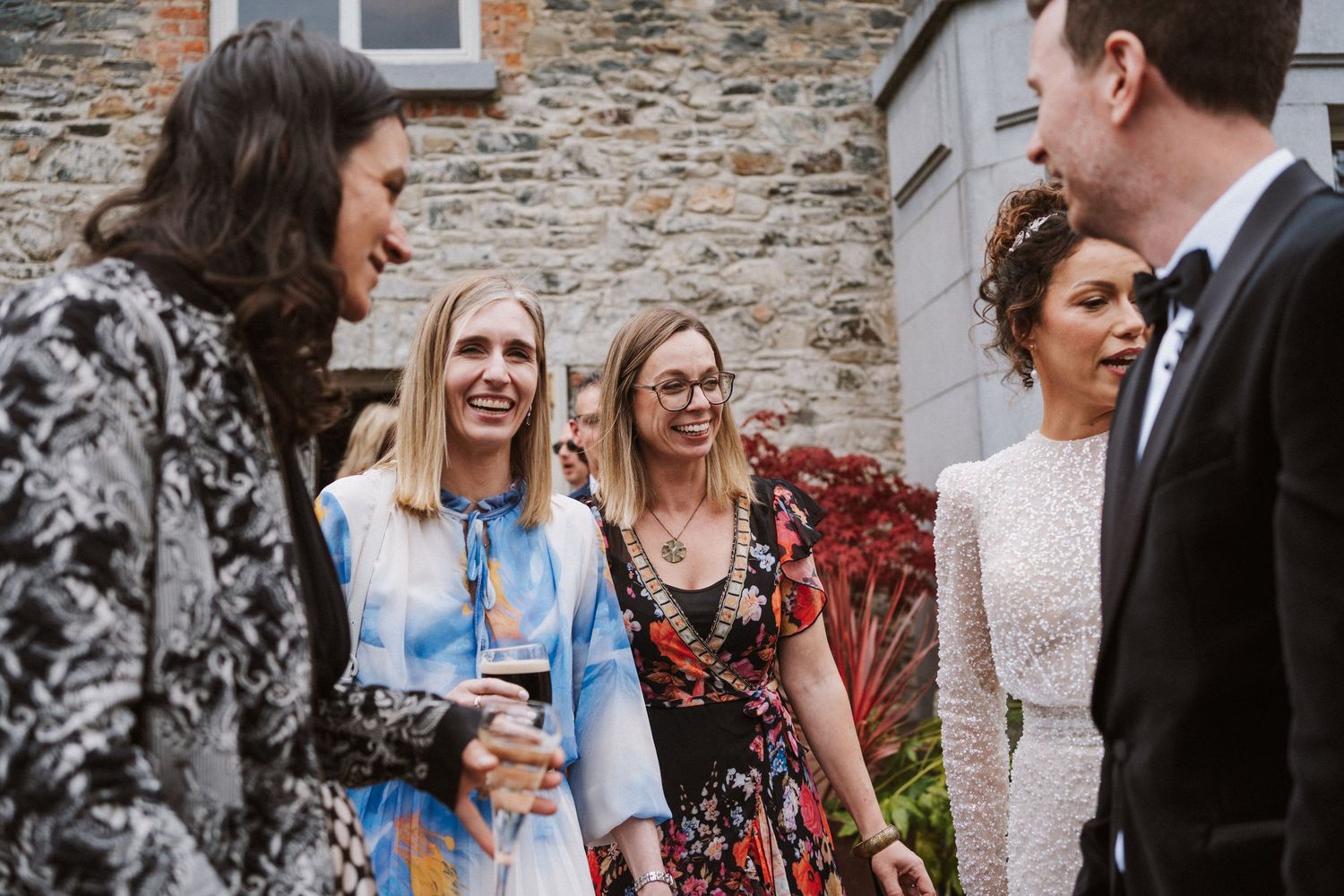 Candid moments of wedding guests laughing and socializing outside a rustic stone building during a celebration.