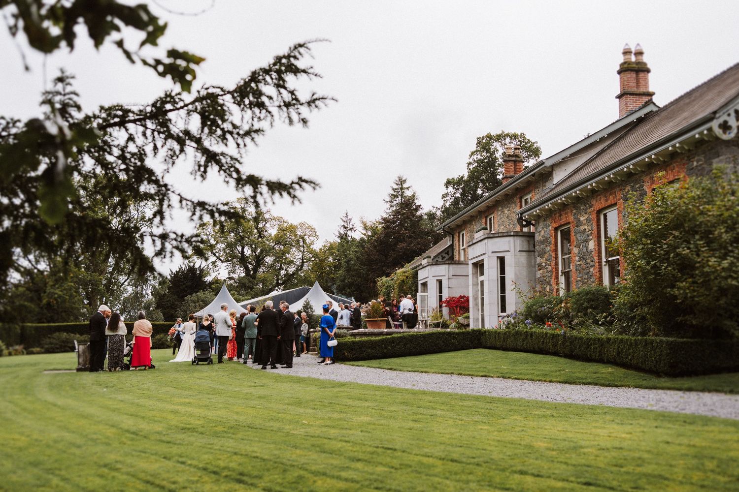 Wedding guests gather on a manicured lawn outside a stone cottage with tents and decorations set up for an outdoor event.