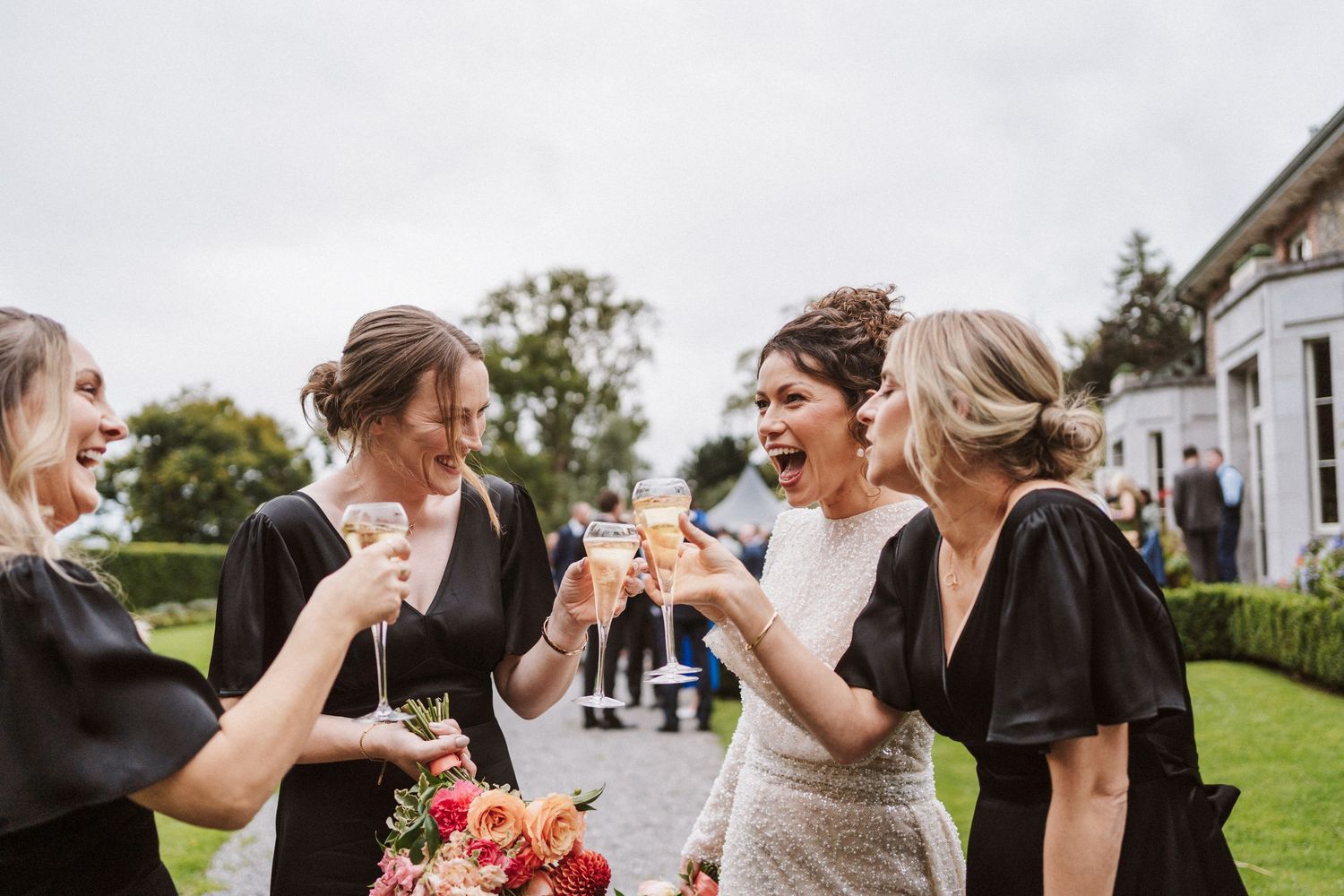 Wedding party dances joyfully in a line on a grassy lawn outside a venue during a celebration.
