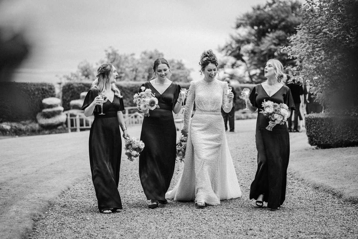 A black and white photo of a wedding party walking together on a garden path, with bridesmaids in dark dresses.