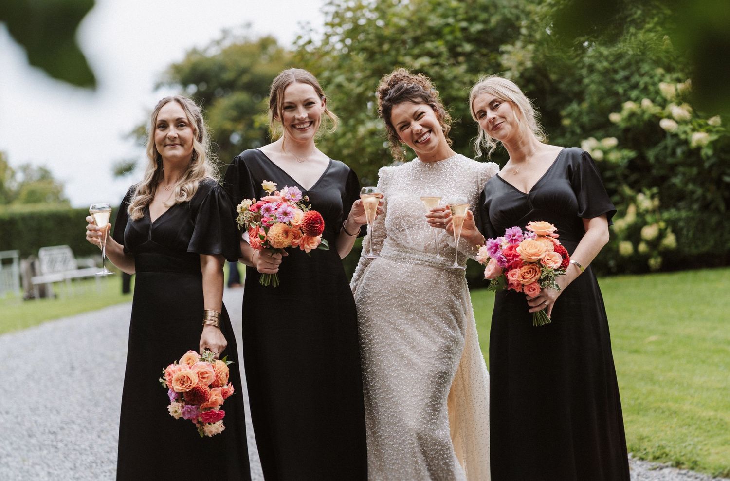 A wedding party in black dresses and white lace gown holding vibrant coral and pink bouquets pose on garden path.