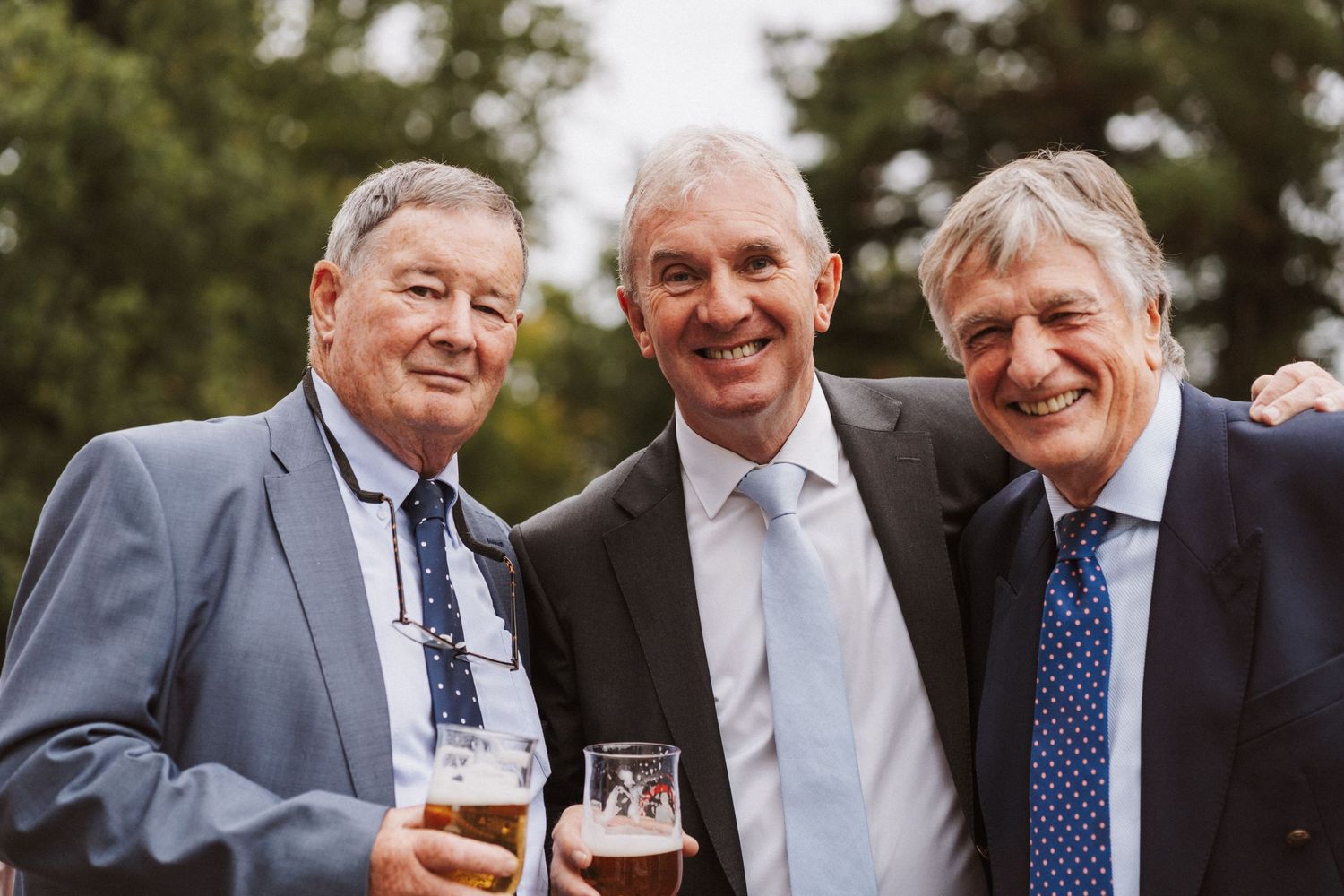 Three men in formal attire sharing laughter and drinks at an outdoor gathering.