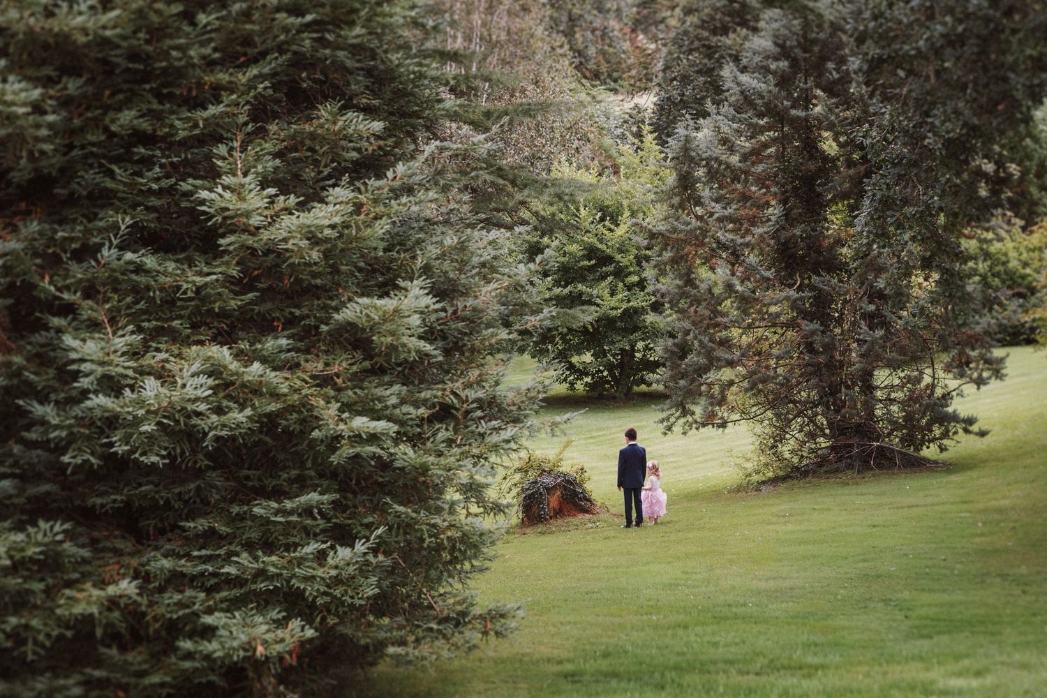 Two figures walk together on a lush green lawn surrounded by tall evergreen trees in a peaceful garden setting.