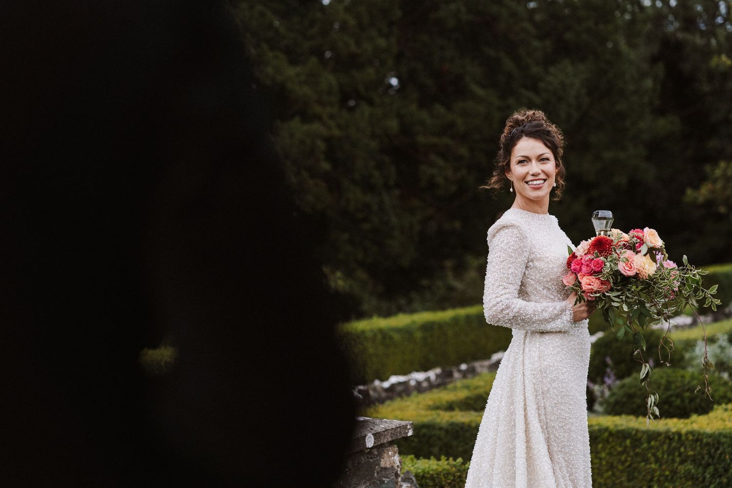 A bride in a vintage-style lace wedding dress holds a vibrant coral bouquet in a garden setting.