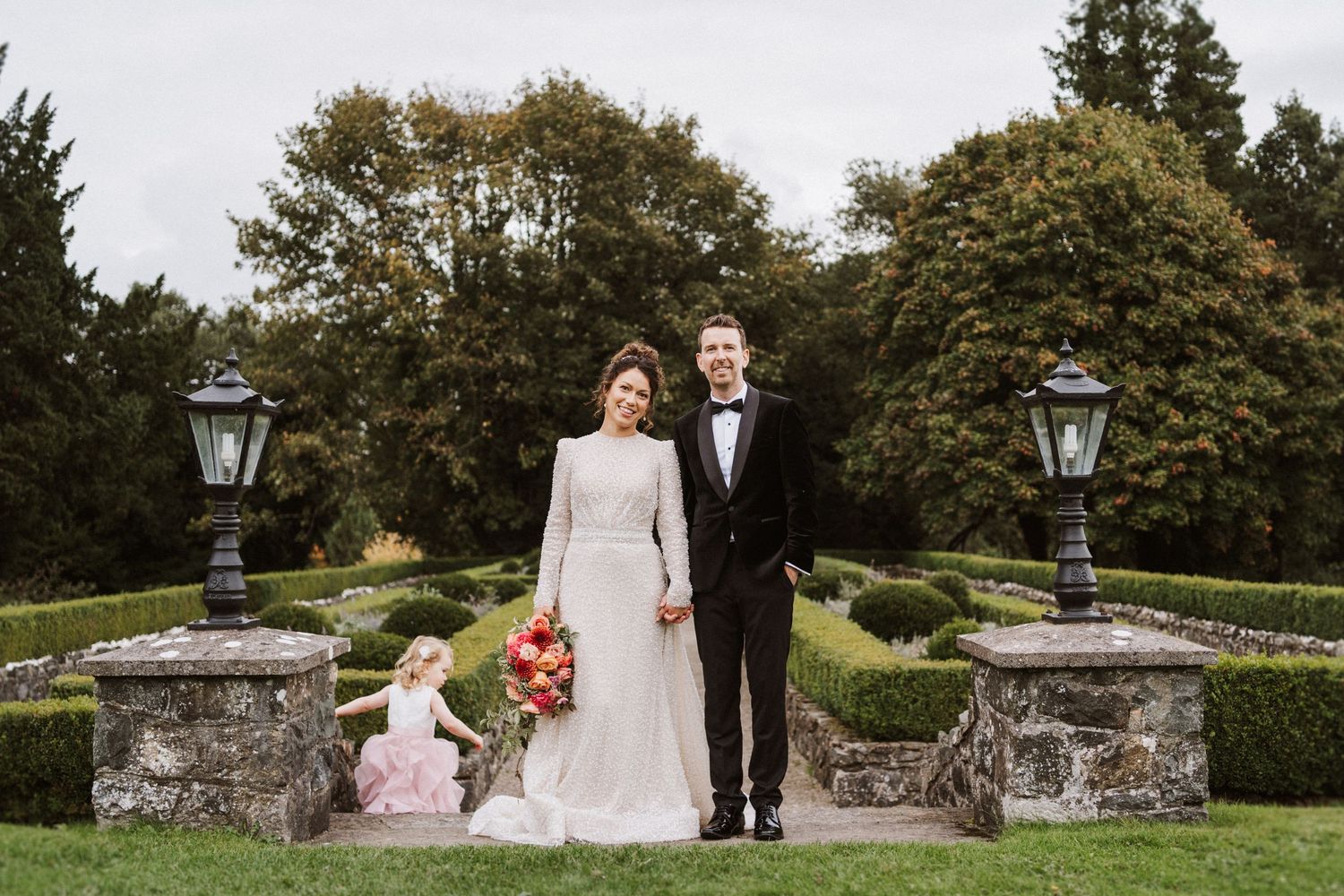 A couple walks through a lush garden setting with stone walls and traditional lamp posts during a wedding celebration.