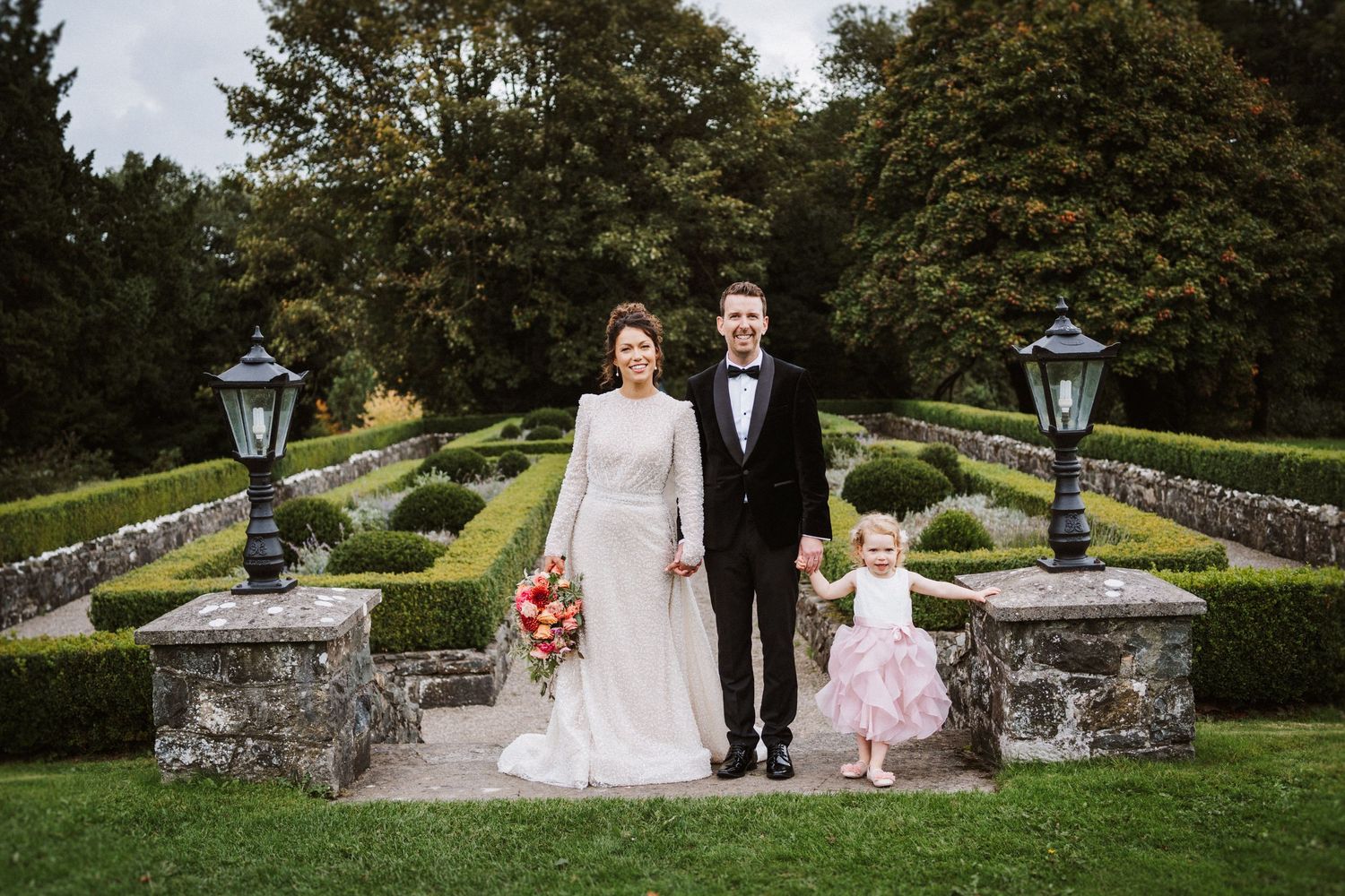 A wedding couple and flower girl pose on stone steps in a formal garden with lanterns and autumn trees.