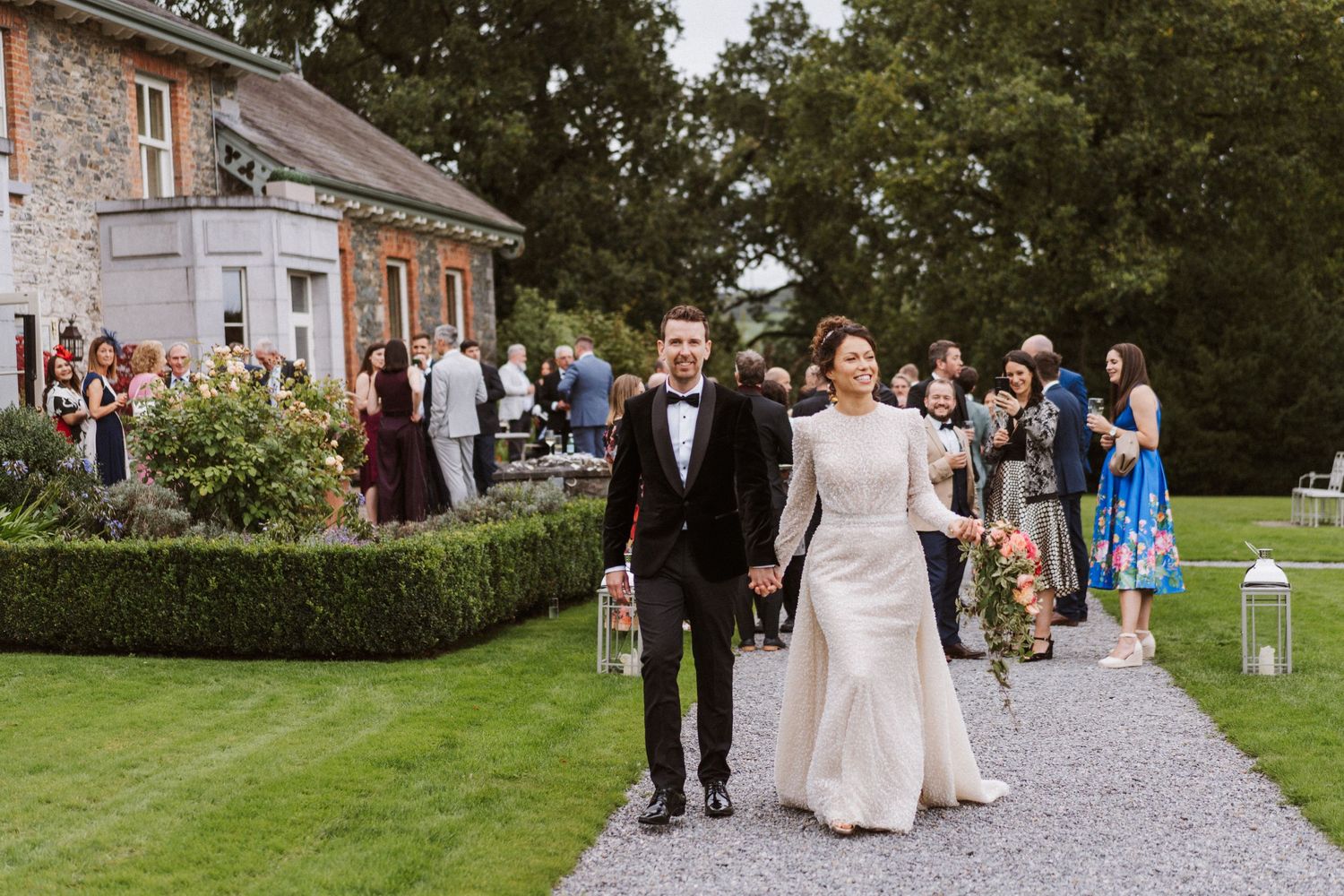 Wedding party guests walk along a stone path outside a historic brick building during an outdoor celebration.