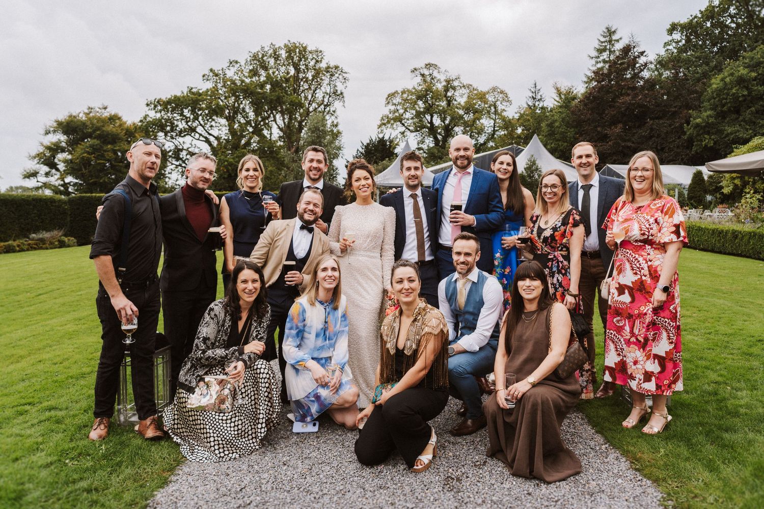 Large group of wedding guests gather on a gravel path for an outdoor group photo at a countryside venue.