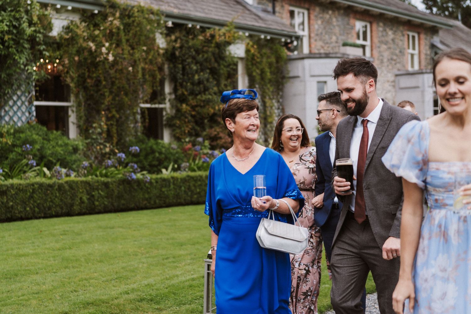 Wedding guests in formal attire gather outdoors on a sunny day near stone buildings and gardens.