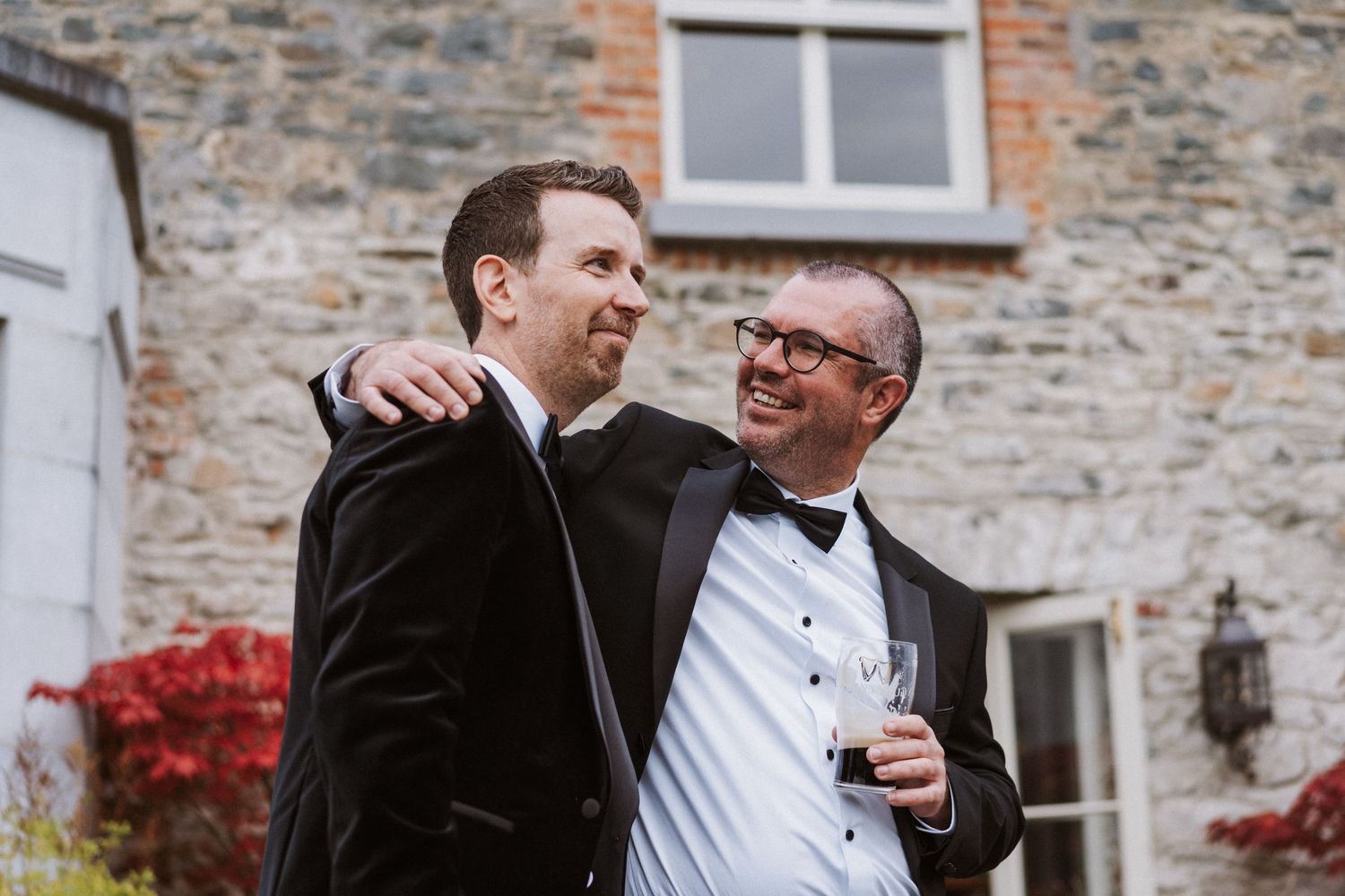 Wedding party laughing together outside stone building with red autumn foliage in background.