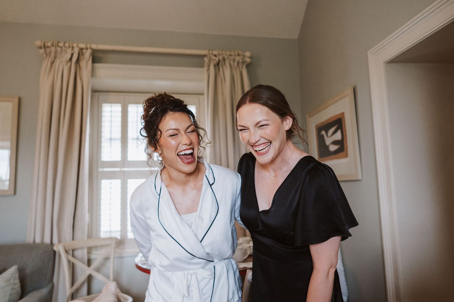 Two people laugh joyfully together in a sunlit room with white curtains and natural decor.