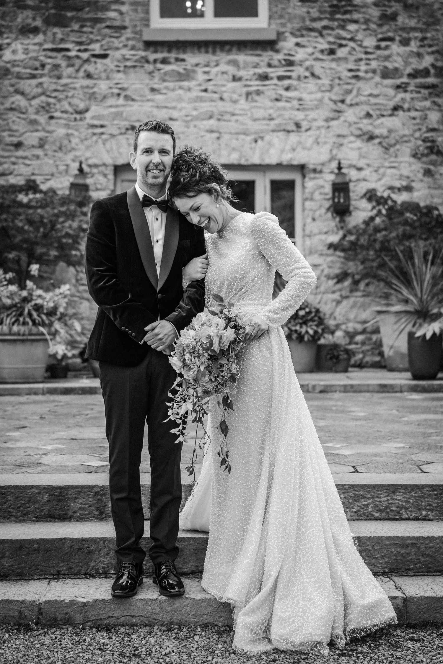 Black and white wedding photo showing couple in formal attire posing outdoors against stone wall backdrop.