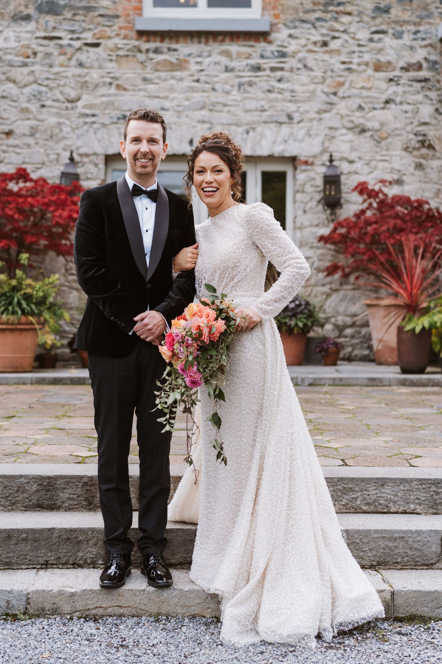 A couple in formal wedding attire poses in front of a rustic stone wall with red maple trees.