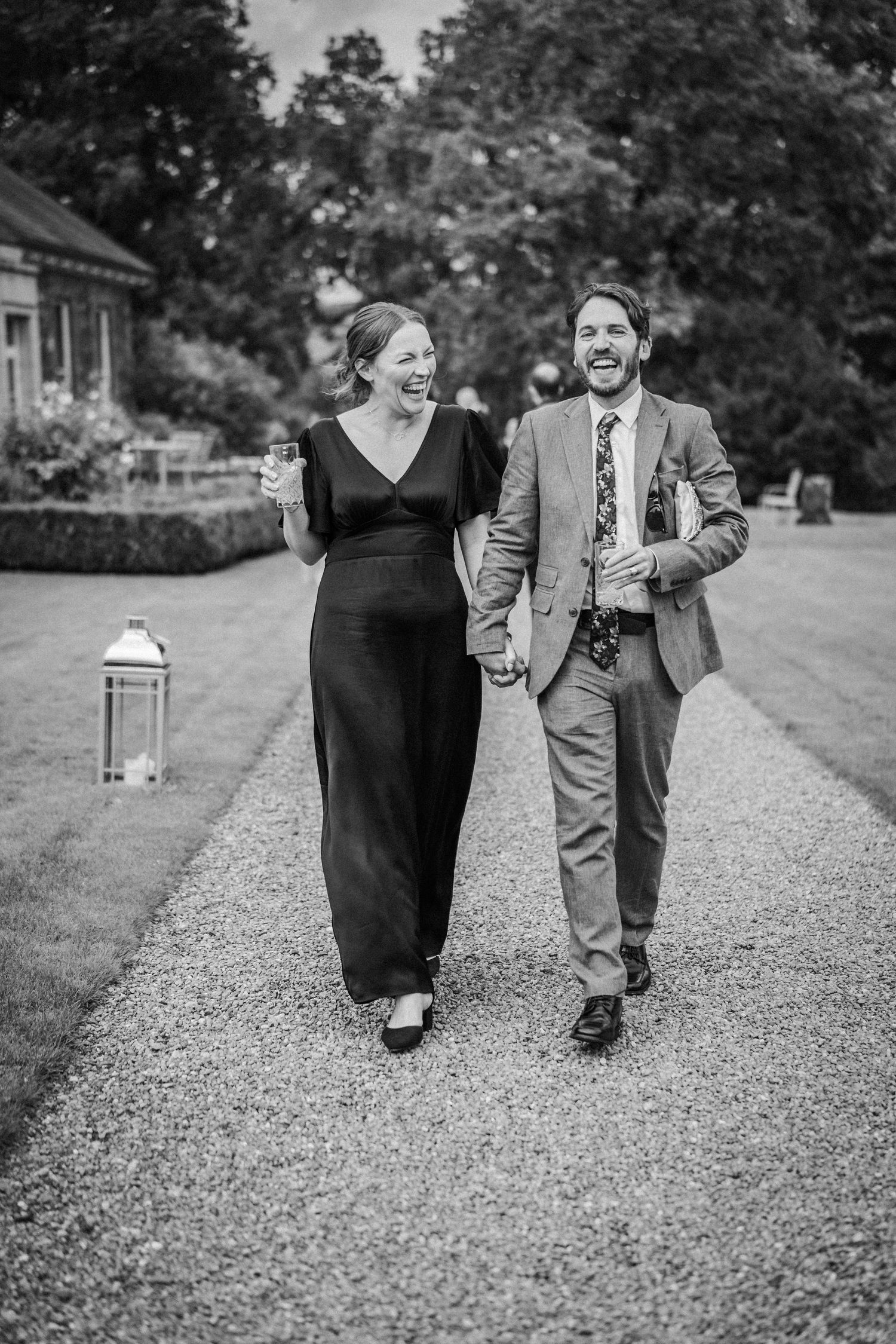 A couple walks together and laughs on a gravel path outside, with the person in a long black dress and suit holding hands.