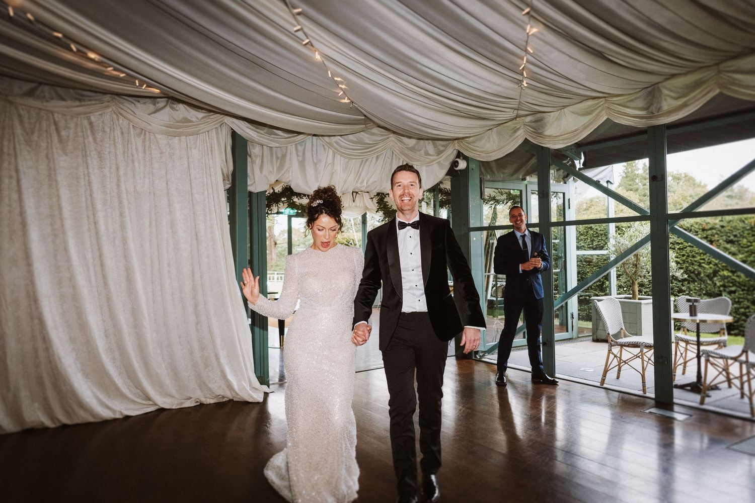 A couple walks together through an elegant venue space with draped white fabric and large windows.