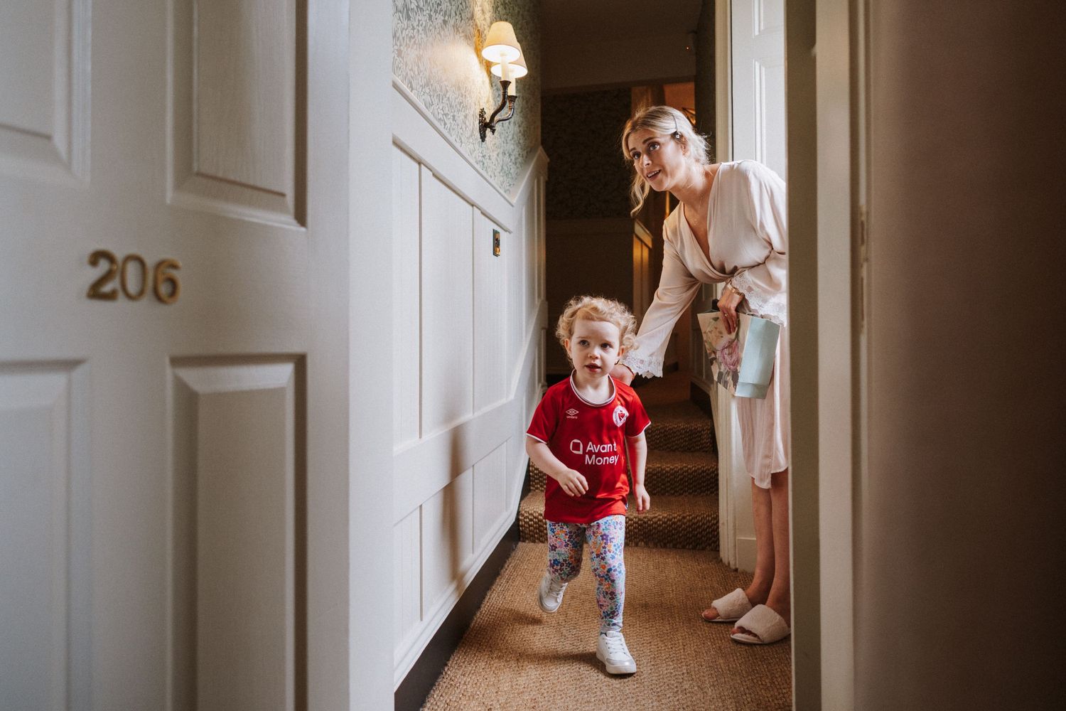 Two children run playfully down a bright hallway while an adult watches from behind.