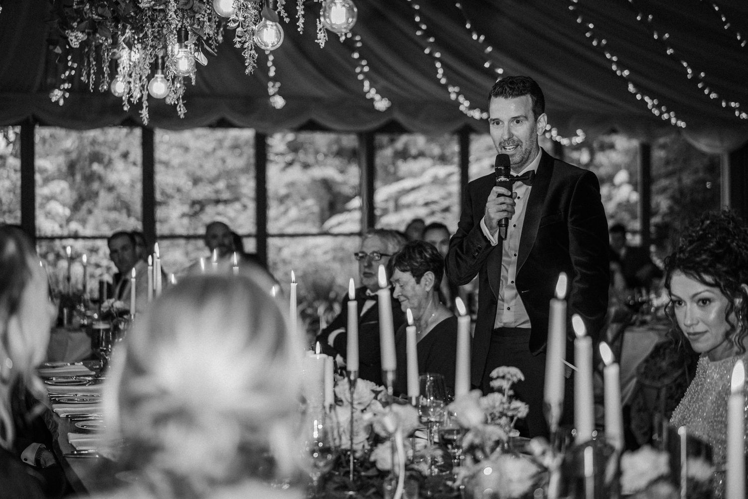 Black and white photo of a wedding reception with string lights and guests mingling under an outdoor tent canopy.