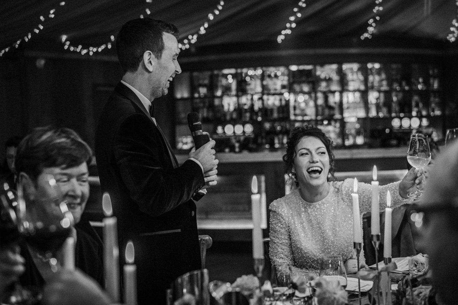Black and white candid wedding reception photo showing guests laughing and enjoying themselves at a dinner table.