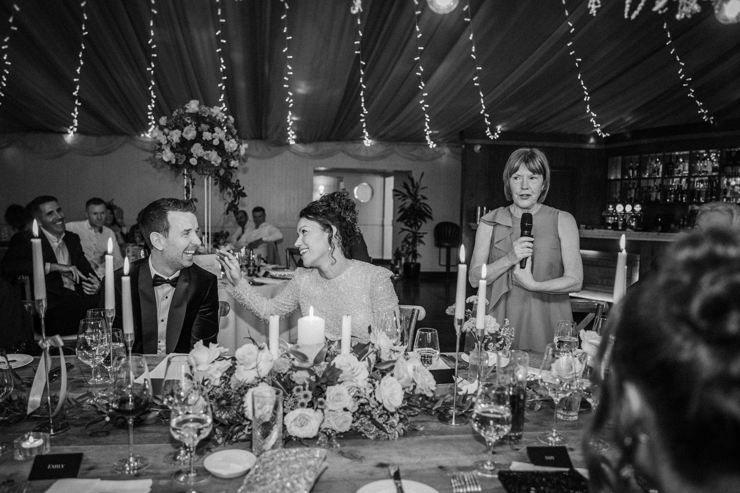 Black and white photo of wedding reception tables set with elegant decor under draped fabric and twinkling lights.