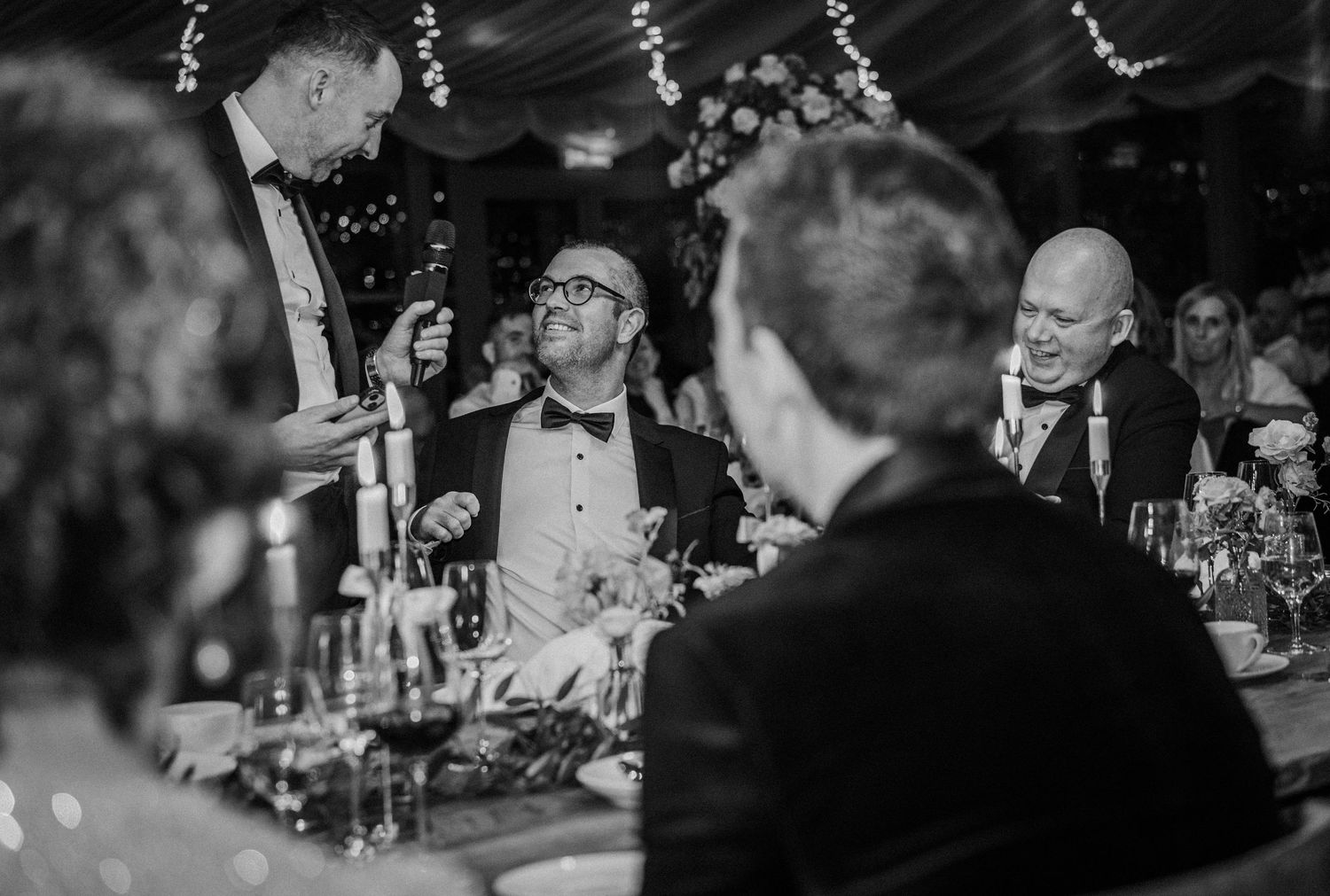 Wedding reception guests seated at tables beneath decorative string lights in black and white photography.