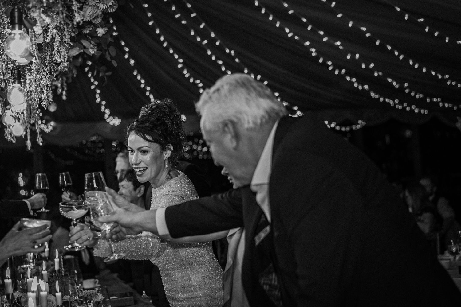 Candid black and white shot of wedding celebration with string lights overhead and guests at dining tables.