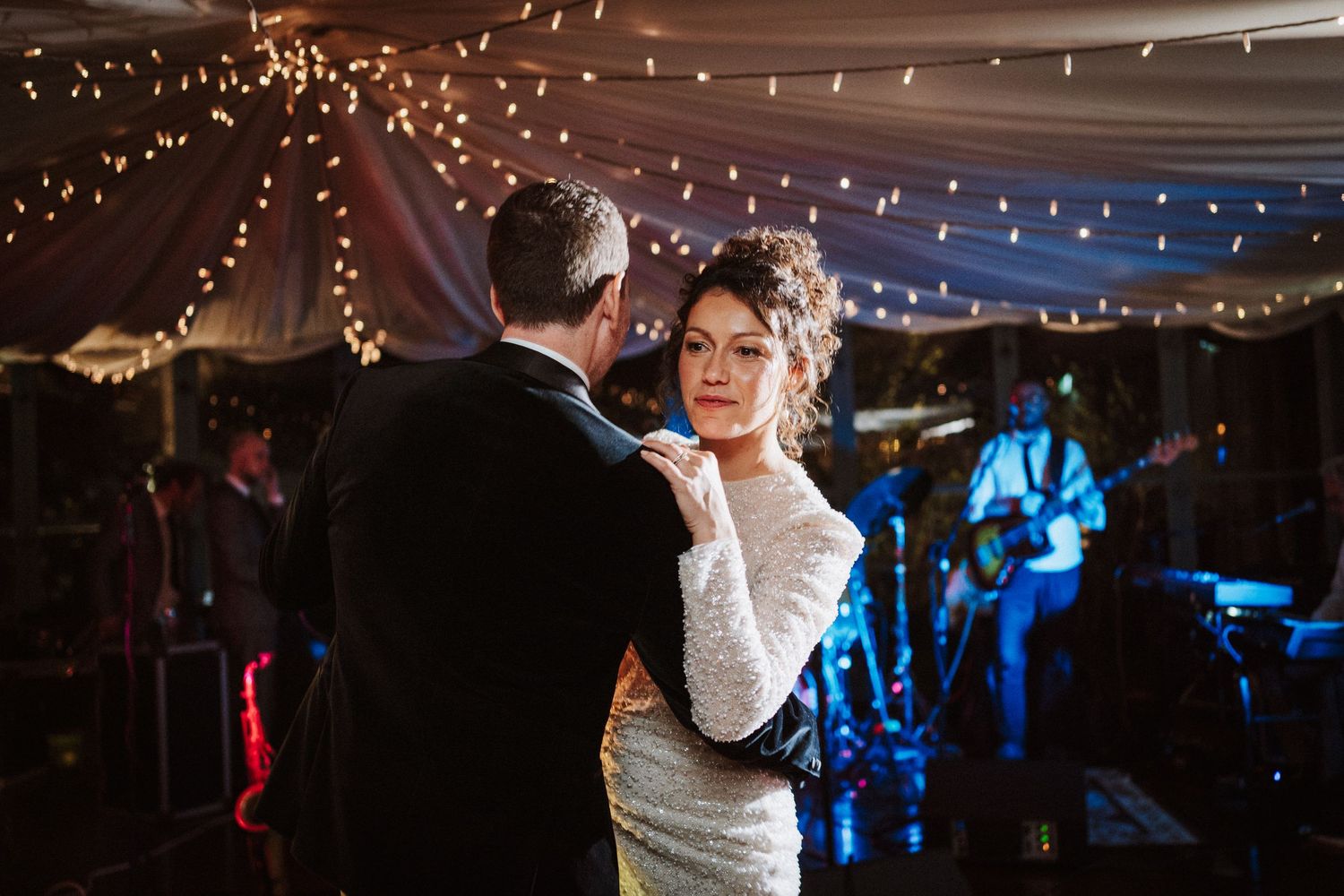 Couple dances intimately under romantic string lights at an evening wedding reception.