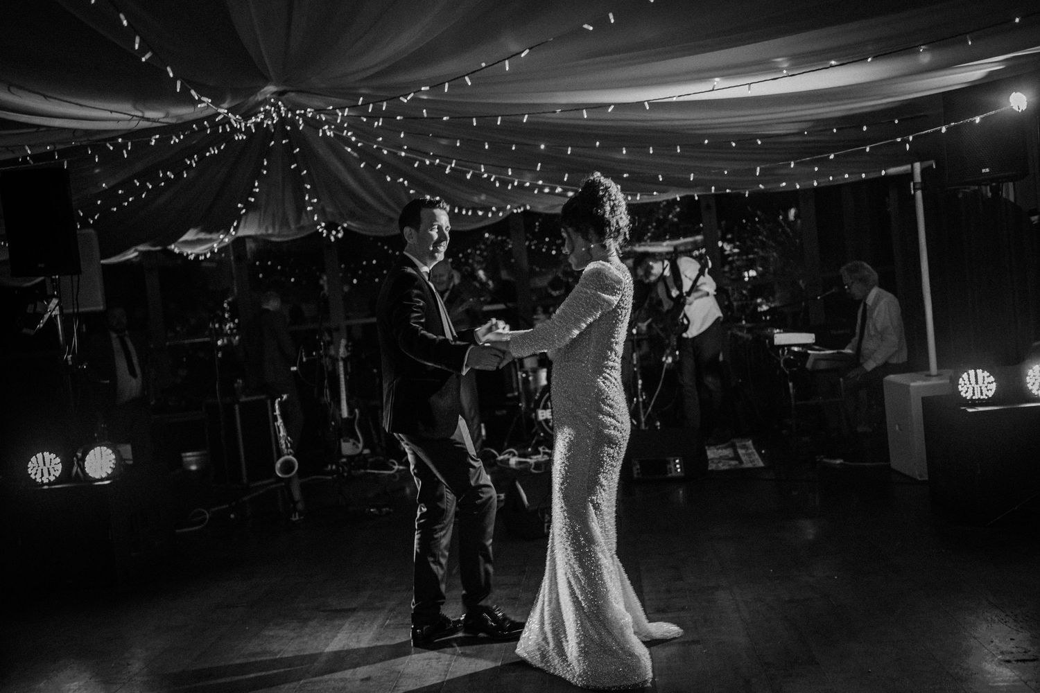 A couple shares their first dance at a wedding reception under twinkling string lights in black and white photography.