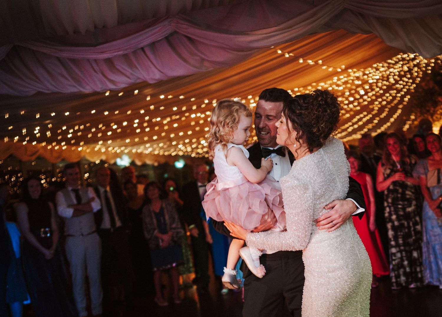 Romantic wedding couple dances under twinkling string lights during their evening reception celebration.