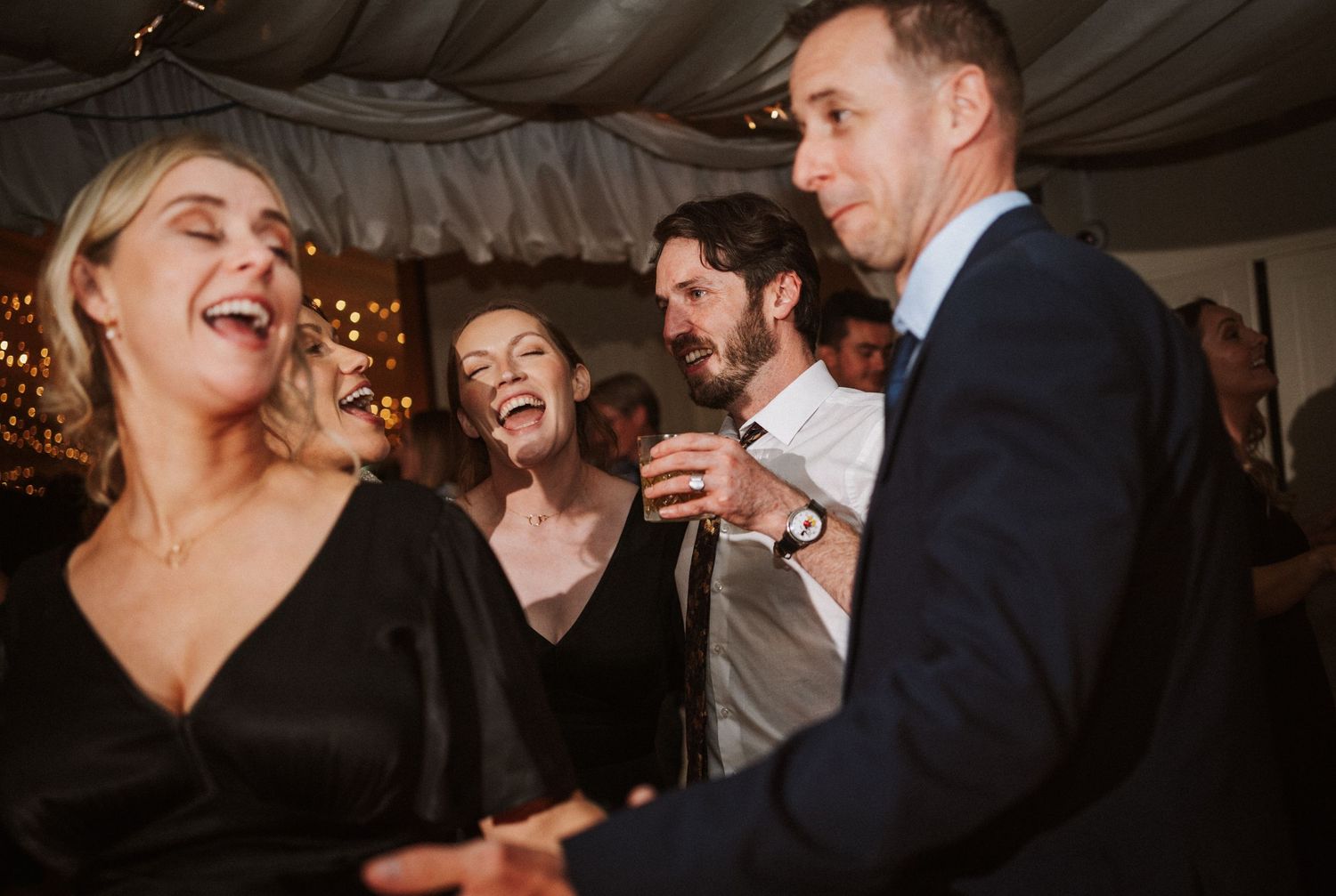 Joyful wedding reception guests dancing and laughing together under a white tent with twinkling lights.