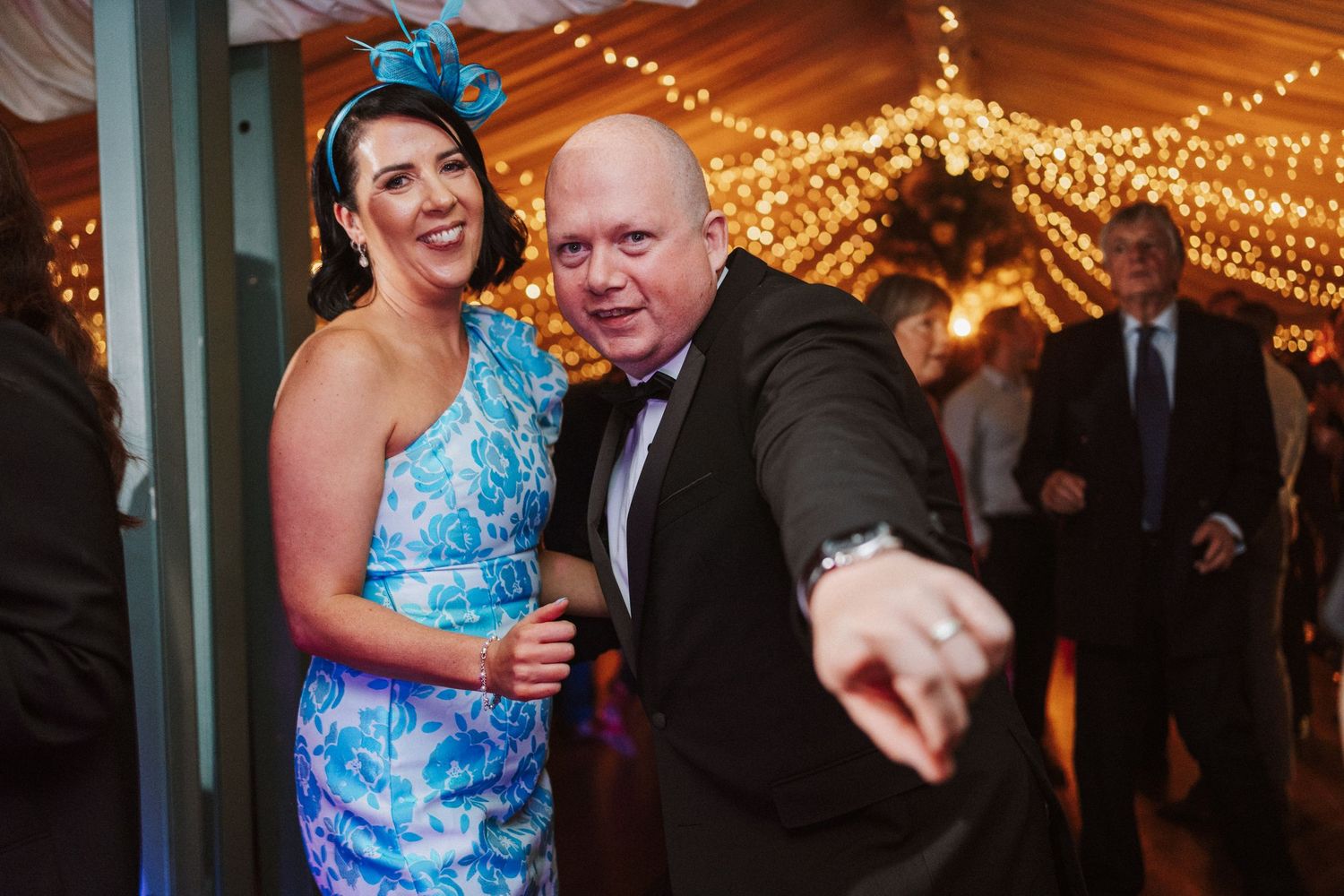 Wedding guests dance and celebrate under warm string lights at an indoor reception venue.