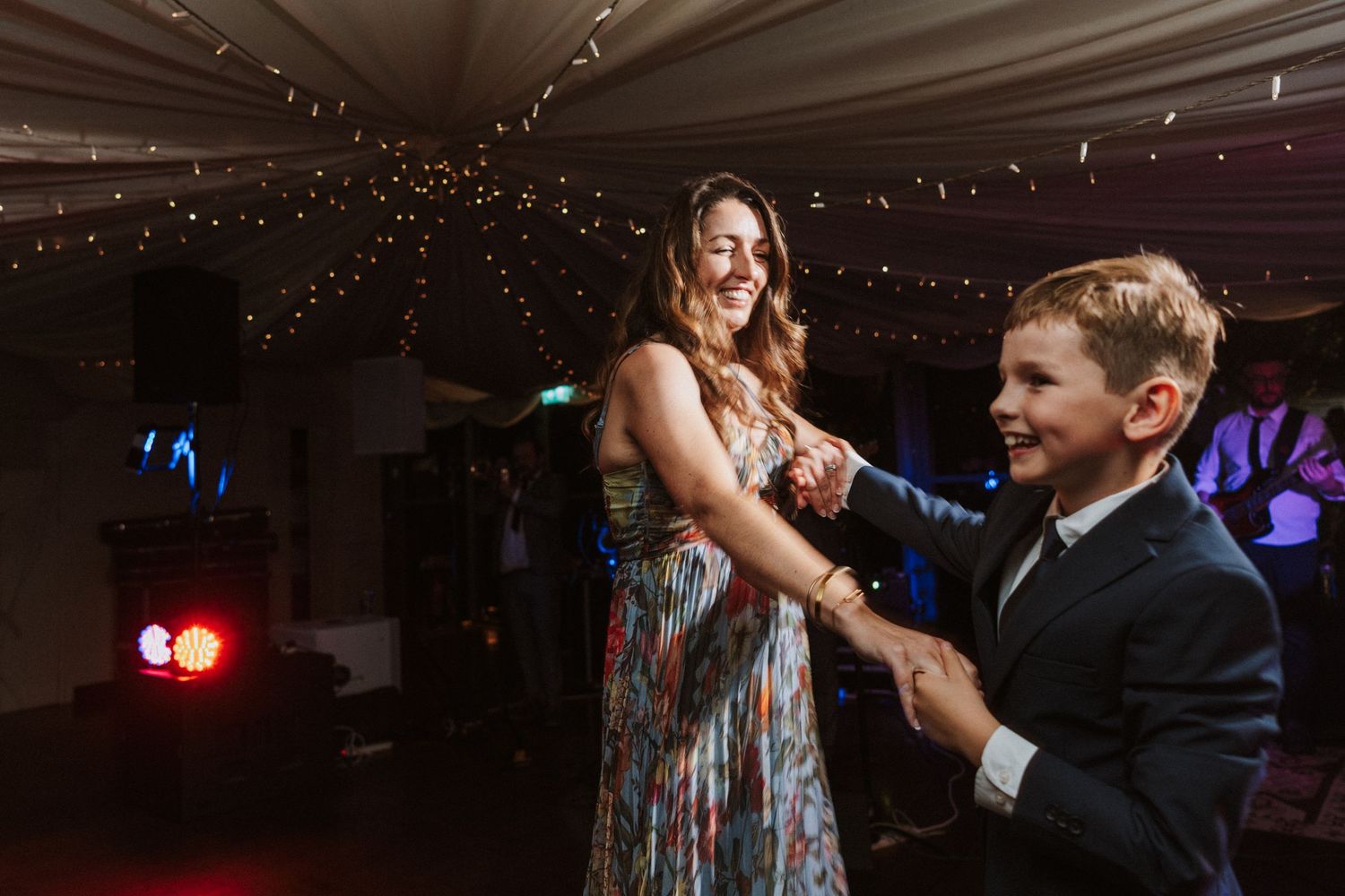 Wedding guests dance and celebrate on a dimly lit dance floor under string lights and draped fabric.