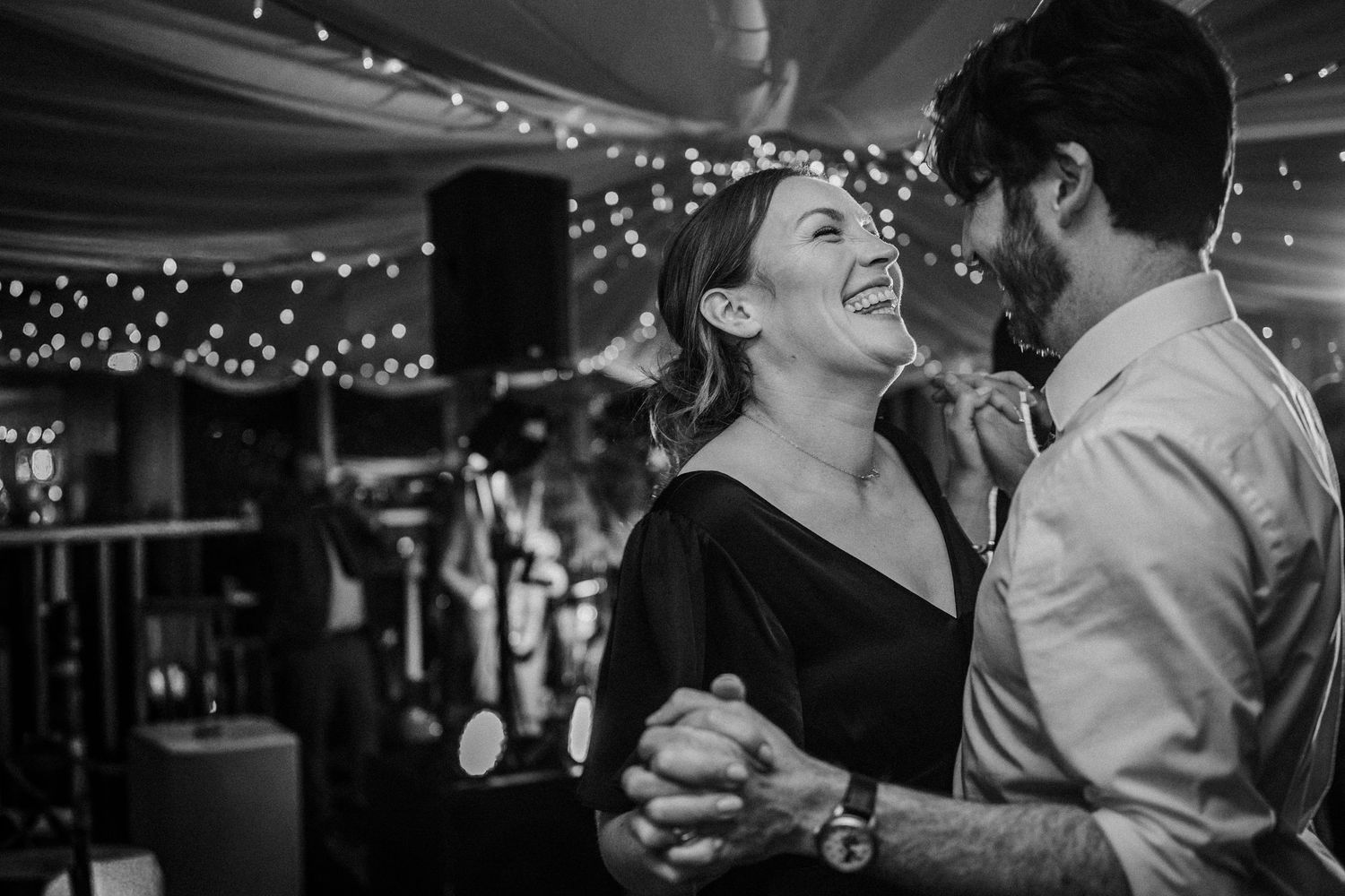Black and white photo of people dancing and celebrating at an elegant indoor wedding reception with twinkling lights.