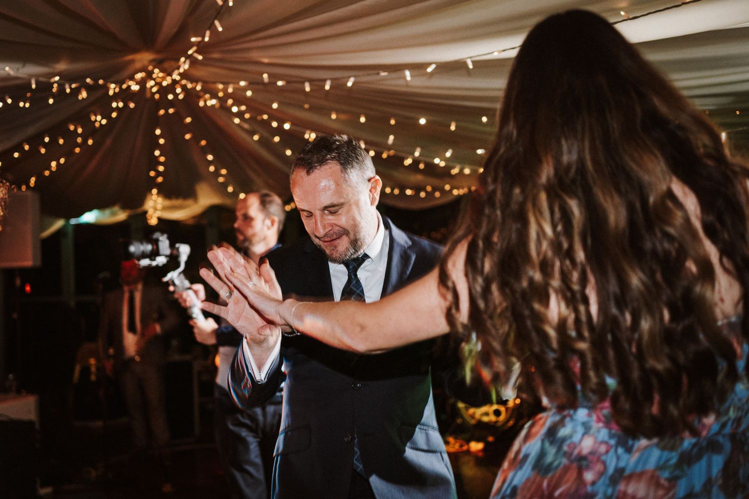 Wedding reception guests dance and celebrate under twinkling lights and white tent draping.