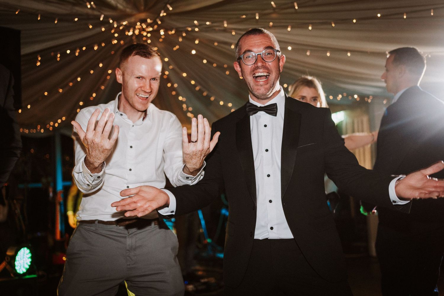 A group of people in formal attire dancing and celebrating at a wedding reception with string lights overhead.